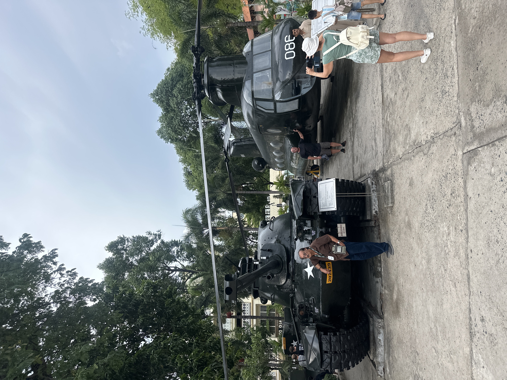 The image captures a scene at a historical site, likely a museum or memorial, in Thủ Đức, Vietnam. An armored vehicle, possibly a tank, is positioned in the foreground, with a man standing next to it and appearing to take a photograph. The tank is painted in dark green and has a white star insignia on its side. A helicopter, dark green with the number "080" painted on its front, is behind the tank. Several people are standing near the helicopter. The ground is concrete, and trees and other vegetation are visible in the background. The sky is visible at the top of the image, indicating it's an outdoor setting.