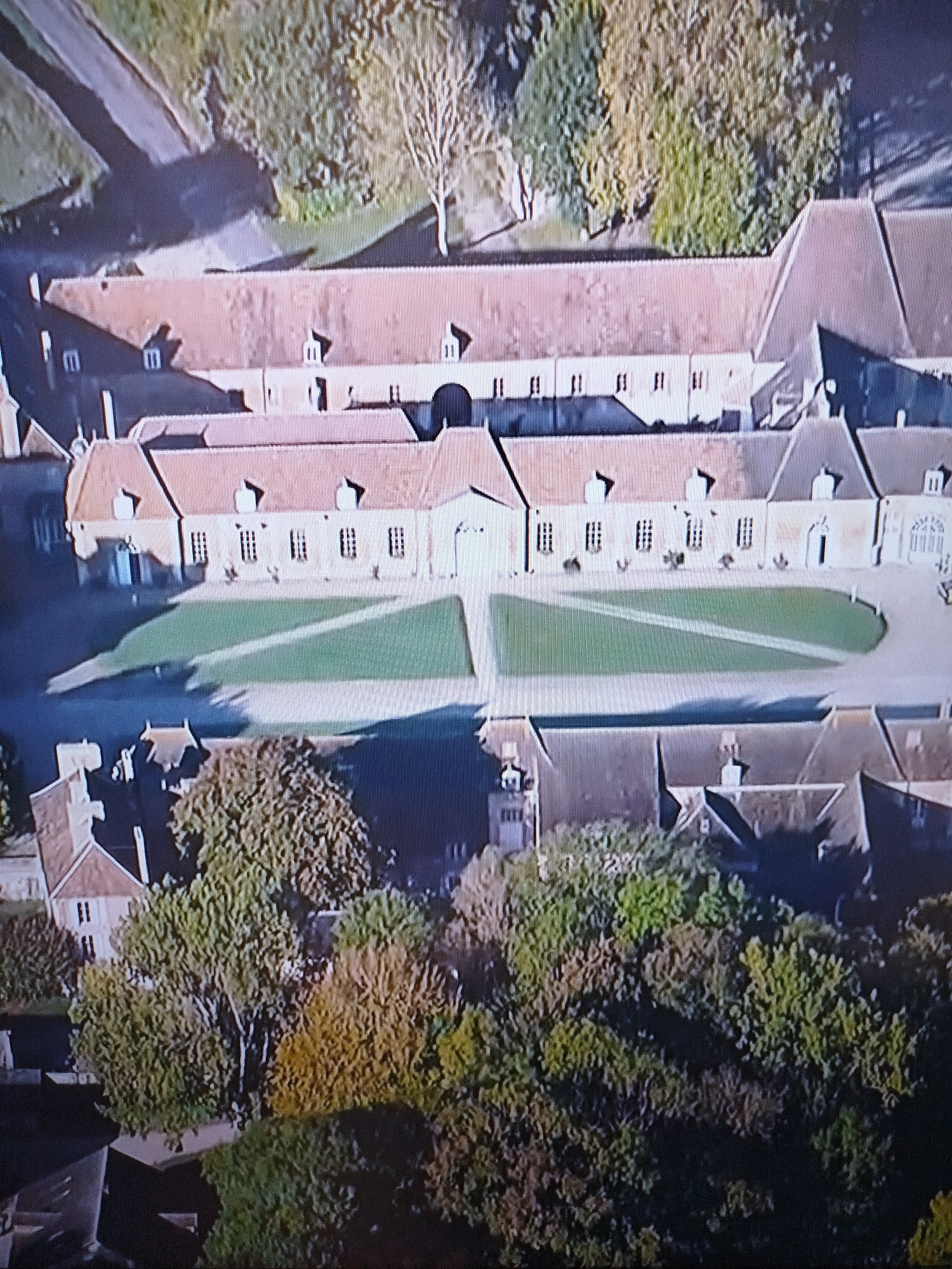 This is an aerial view of a large, sprawling estate or complex, likely a former monastery or aristocratic residence, located in Naberezhnye Chelny, Russia. The architecture suggests European influences, with red-tiled roofs and symmetrical building layouts. The main building appears to be a long, two-story structure with numerous windows and dormer roofs. In front of this building, a large, manicured lawn forms a central courtyard, with pathways cutting across it. Surrounding the central complex are smaller buildings, outbuildings, and dense greenery, including trees with autumnal foliage. The overall impression is one of historical significance and a peaceful, rural setting.