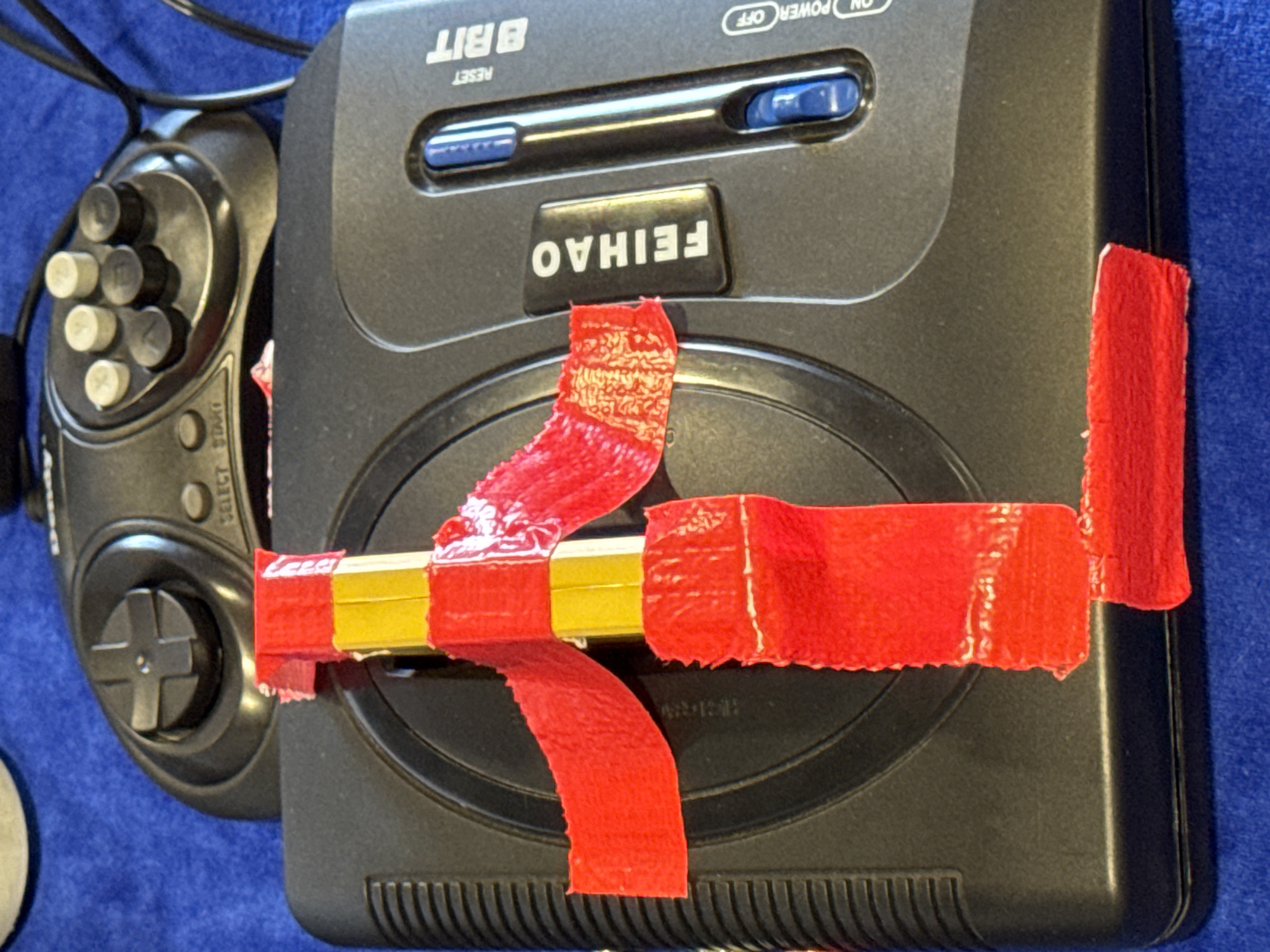 The image shows a close-up of a black Sega Genesis video game console with red and yellow tape wrapped around it. A black video game controller with a directional pad and buttons is partially visible in the upper portion of the image. The console and controller are placed on a blue fabric surface. The text "FEIHAO" is visible on a black button on the console, and "8 BIT" is printed on the console as well. There are also labels for "RESET", "ON", and "OFF" near switches. The scene is static and appears to be indoors, with no discernible city or location information present.