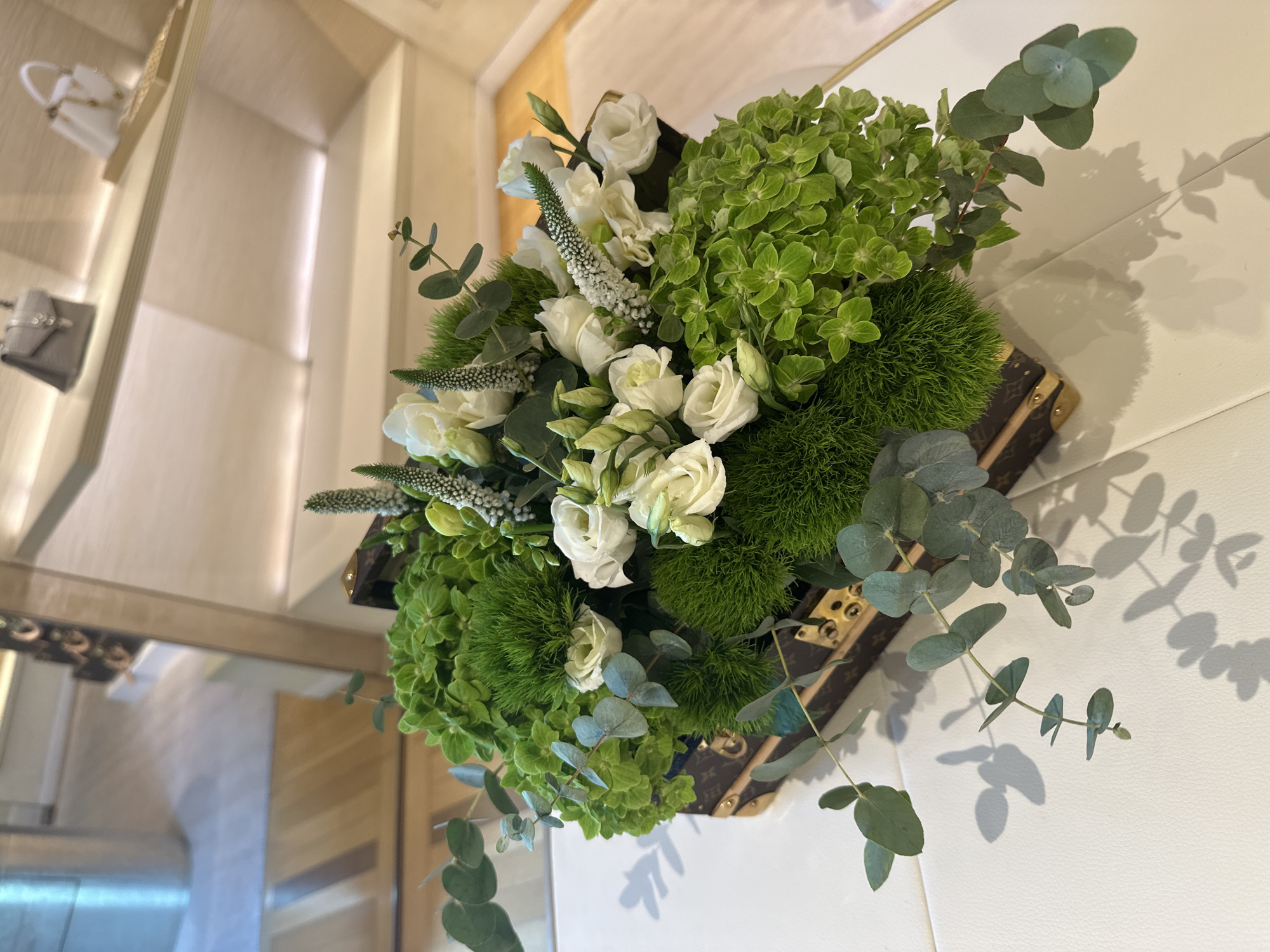 The media features a sophisticated floral arrangement centered in an indoor setting. The bouquet is a harmonious mix of white and green elements, comprising elegant white roses or similar white clustered blooms, lush light green hydrangeas, textured spherical green Dianthus 'Green Trick' or a similar plant, and graceful sprigs of eucalyptus.
This vibrant assembly is presented in a distinctive rectangular container that strongly resembles a vintage Louis Vuitton trunk. The container is covered in material featuring the iconic Louis Vuitton monogram pattern and is accentuated with brass-colored corner protectors and latches, signaling luxury and brand identity.
The background, subtly out of focus, reveals a bright, upscale interior. A light-colored, likely white or cream, textured surface stretches across the lower and right parts of the image, where soft shadows of the eucalyptus leaves are visibly cast. Reflections in the upper left section suggest a well-appointed display area with shelves holding various handbags or purses, including a white and a grey one, indicating a retail or showroom environment. The lighting is warm and diffused, contributing to a refined ambiance.
There is no human activity or event actively taking place; the image captures a static, decorative display designed to enhance the sophisticated interior. The overall impression is one of elegance and high-end luxury. This kind of arrangement and setting is characteristic of luxury establishments, fitting perfectly within the context of a high-end store or hotel in a city like Madrid, Spain. The visible text consists of the Louis Vuitton monogram on the container.