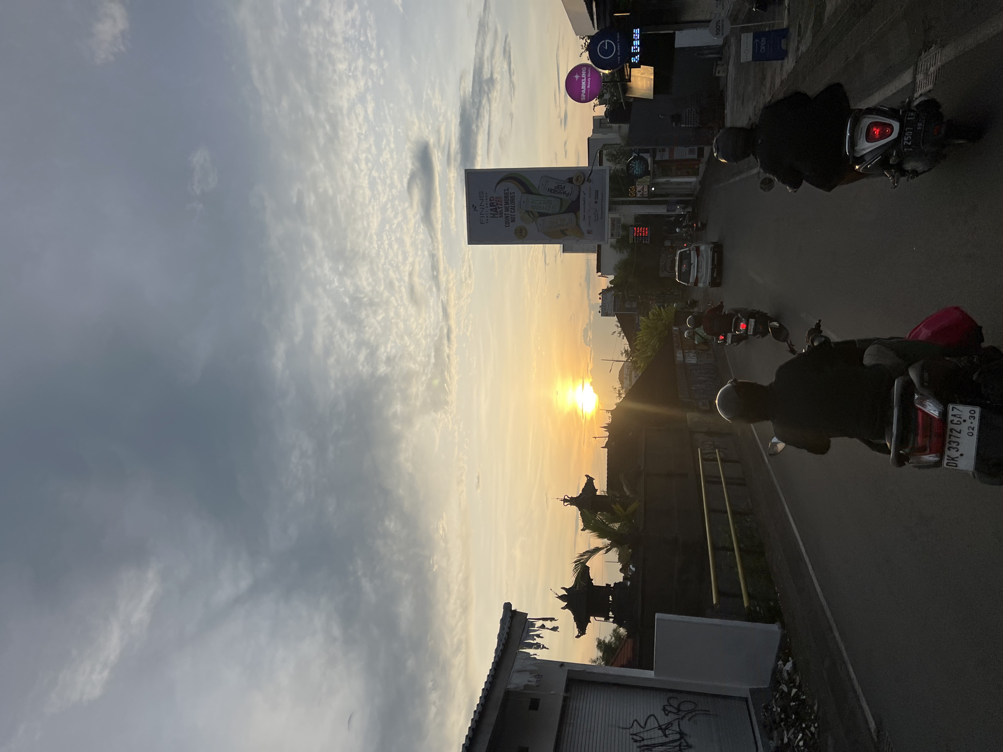Here's a concise description of the image:
The image shows a street scene at sunset in North Kuta, Indonesia. The sun is setting in the distance, casting a golden glow over the scene. In the foreground, there are several people riding motorcycles. A large billboard advertising "FINNS PARTY SHINE HARD SELTZER" is visible. Street signs for "Sparkling" can be seen near a money exchange. A building is visible on the bottom left of the frame, with graffiti visible on it.