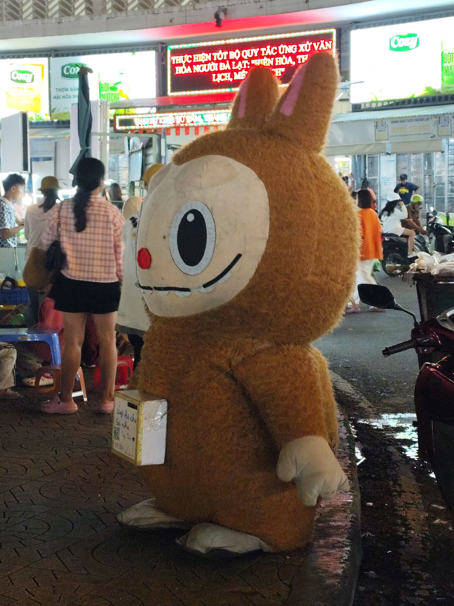 The image depicts a person in a large brown rabbit costume with a white face mask standing on a wet, patterned sidewalk in Da Lat, Vietnam. The costume has a donation box attached to its side. Behind the costumed figure, there are several people, some sitting on small stools or standing. There is a woman with a plaid shirt and short black skirt standing in the background. In the distance, there are motorbikes and shops with lit-up signs, including ones that read "Cozy" and "BOT". The setting appears to be a bustling street at night, possibly near a market in Da Lat.