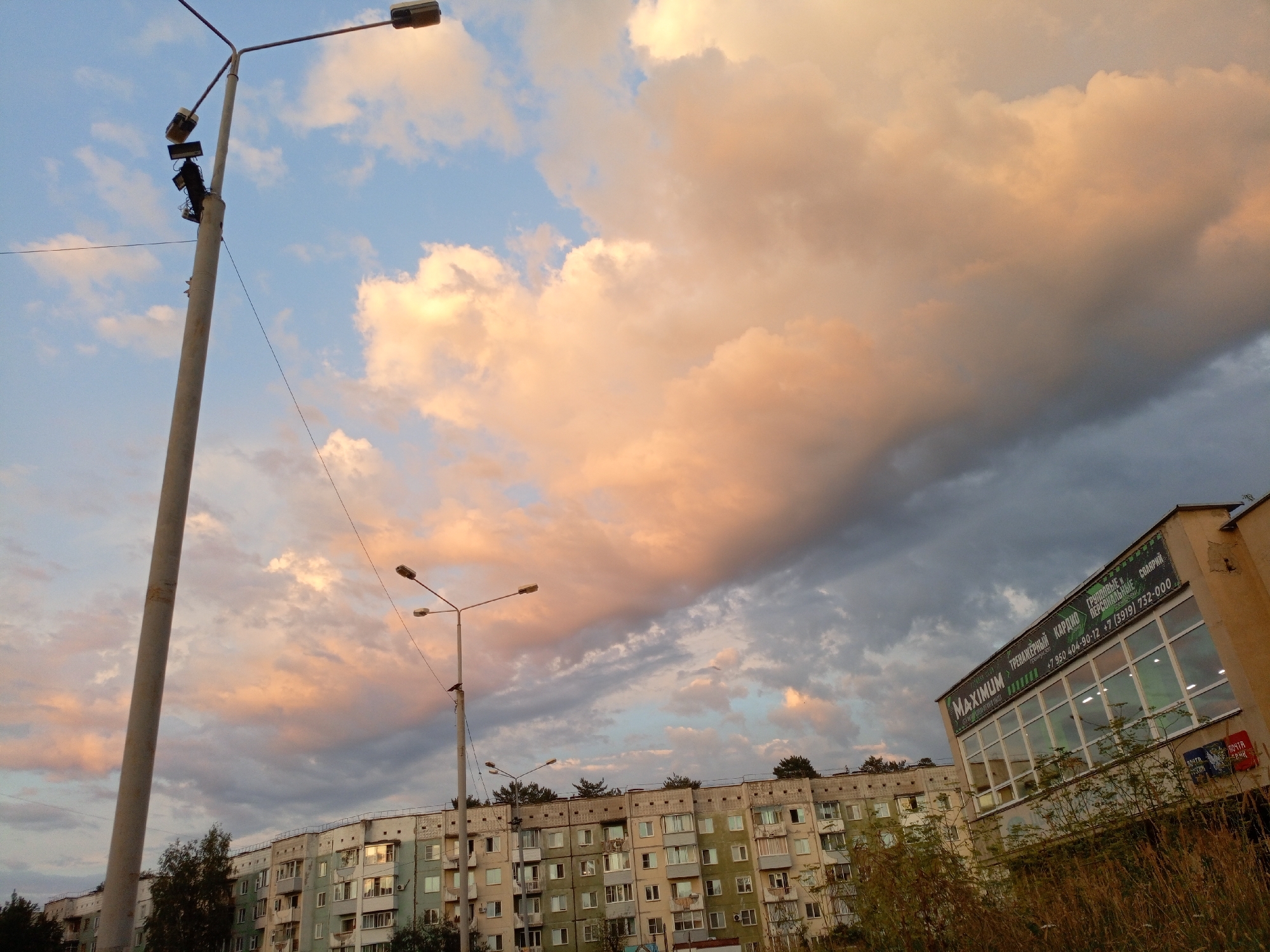 The image depicts a street scene in Zheleznogorsk, Russia, at dusk. The sky is filled with a mix of pale blue and dramatic clouds illuminated with hues of pink and orange from the setting sun, contrasting with darker grey clouds. In the foreground, a tall, grey concrete lamppost with multiple light fixtures stands to the left. Further back, another lamppost with two lights is visible in front of a long, multi-story apartment building. The building is painted in various shades of light green and beige, with numerous windows and balconies. To the right, a building with a large sign advertising "MAXIMUM" and what appears to be a gym or fitness center is prominent, with large reflective windows. The overall impression is of a residential area on the outskirts of a city under a striking sky.