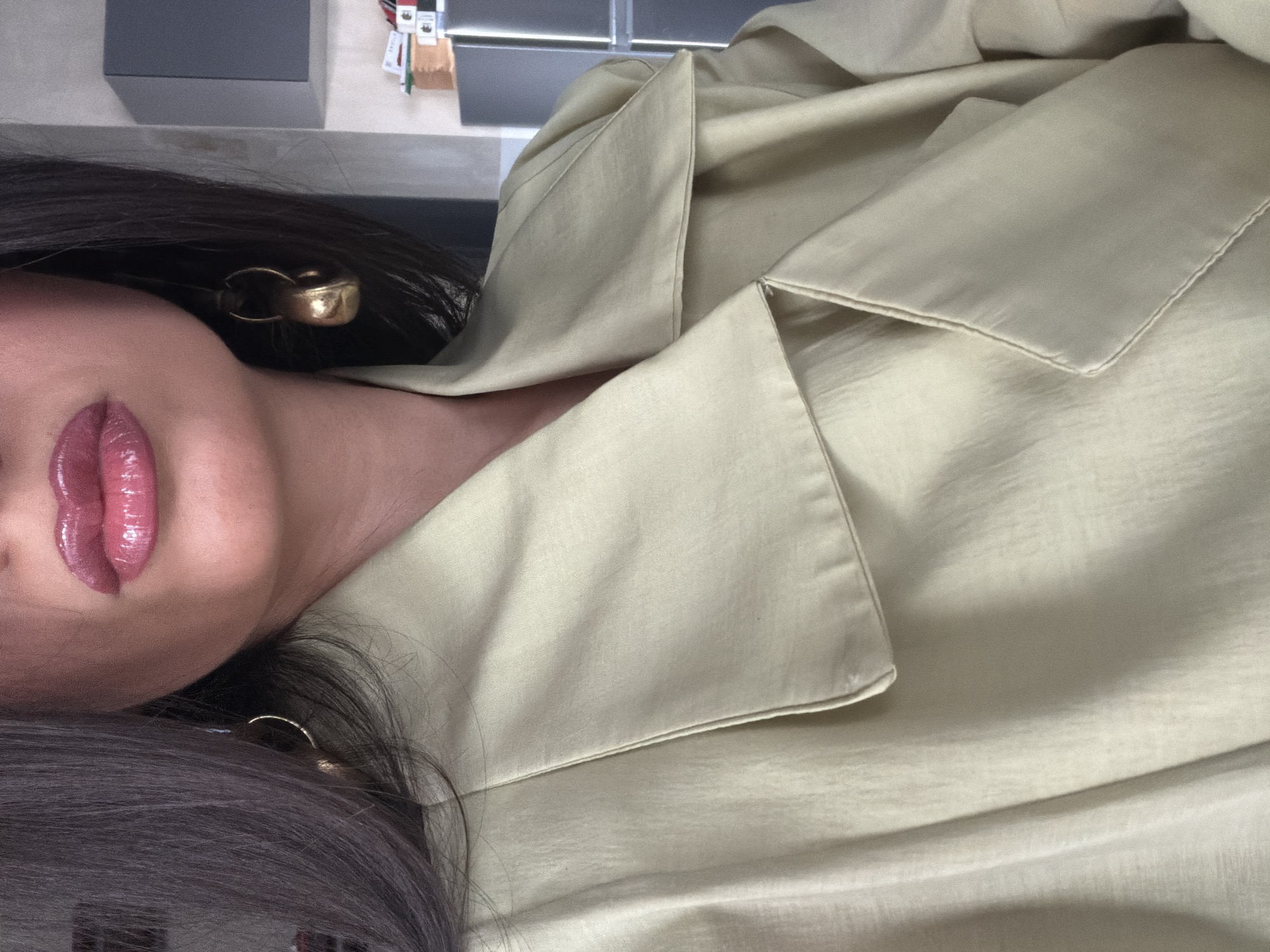 The image is a close-up, sideways-oriented portrait of a person, likely a woman, showcasing her upper body, face, and clothing indoors. The subject has dark hair and wears glossy, plump pink lipstick. A prominent golden-colored, chunky earring is visible on her ear.
She is dressed in a light-colored garment, possibly a shirt or blazer, in a soft, pale yellow or cream hue with a subtle sheen, suggesting a material such as silk or satin. The collar and lapel of the garment are clearly visible, displaying fine stitching details.
The background is intentionally blurred, featuring indistinct rectangular shapes in dark and light grey tones, suggesting an indoor setting like a modern room or commercial space. Small, colorful items, possibly labels or papers, are faintly discernible in the upper right background.
The individual appears to be posing for the camera, with the focus primarily on her makeup, jewelry, and fashion. Her lips are gently parted, conveying a relaxed or contemplative expression. No specific activity beyond posing is evident. There is no visible text in the image. While the location context is Yerevan, Armenia, this is not visually identifiable from the image itself.