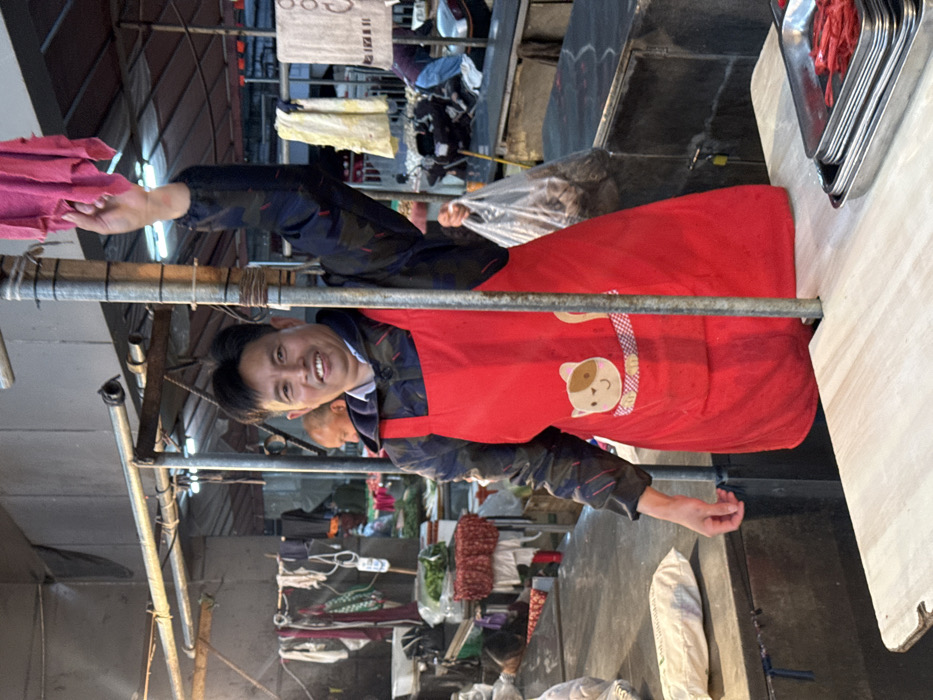 The image captures a market scene in Jiangjin, China. A male individual of East Asian descent, appearing middle-aged, is positioned centrally behind a market stall counter. He wears a dark blue long-sleeved jacket over a light-colored collared shirt, and a red apron adorned with a cartoon cat face on its front pocket. The man is smiling and looking directly at the camera. His left arm is raised, holding a pink textile cloth draped over a vertical metal pole that stands in front of the counter. His right arm is partially visible below the elbow.
In the immediate foreground, to the right on a light-colored wooden surface, are stacked silver metal trays; the topmost tray contains multiple thin, red, elongated items. To the left, on a darker counter surface, a white package is visible. The background reveals additional market activity: a blurred individual is partially visible behind the main subject's left shoulder. Further back on the right, another person wearing a blue garment is seen bending over a counter. Various items, including green vegetables, hanging cloths, and a white plastic bag, populate the surrounding market stalls. A scooter or motorcycle is parked in the mid-ground behind the market counters on the right. The environment is illuminated by overhead artificial lighting, consistent with an indoor or covered market structure.