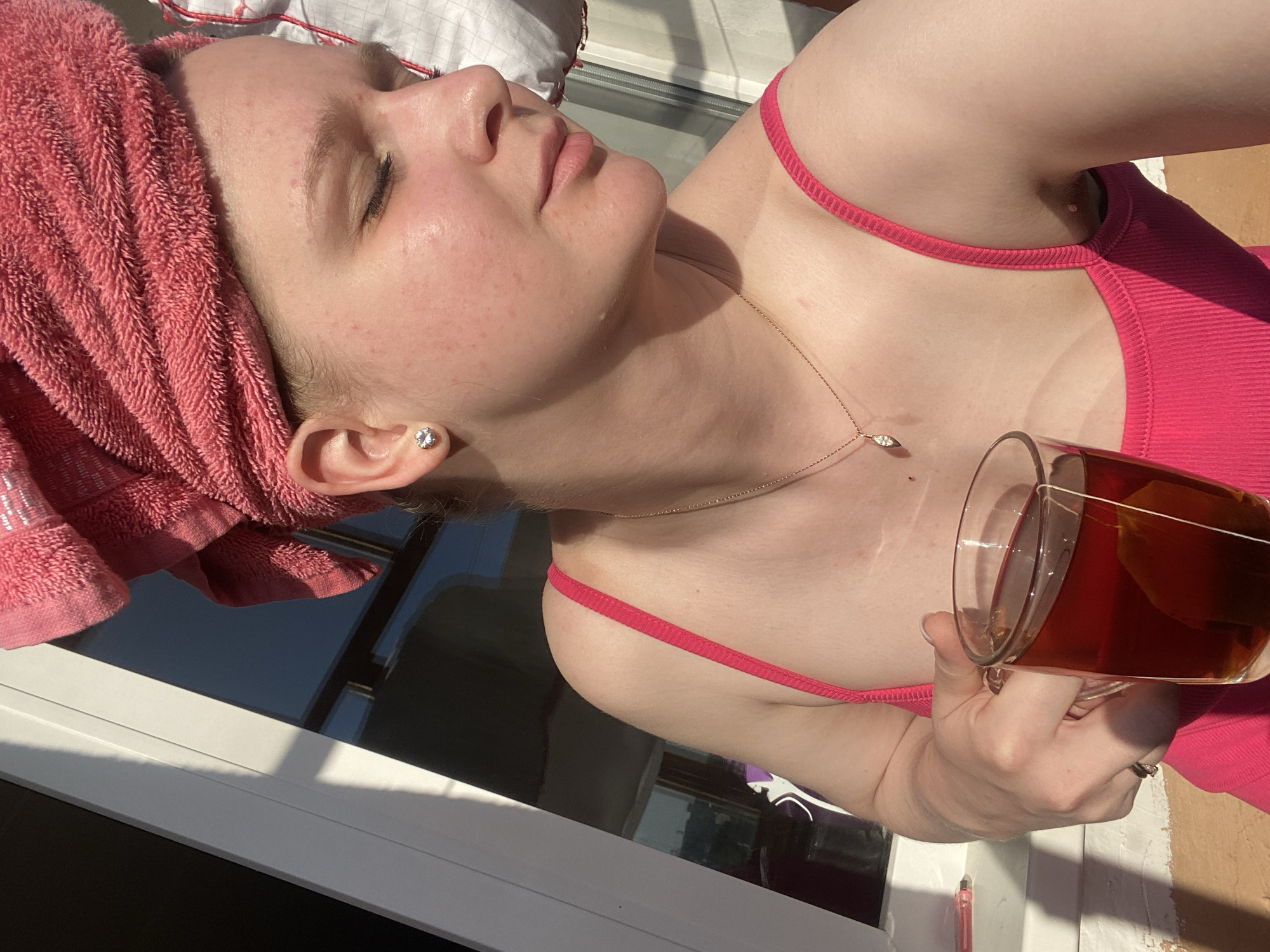The image shows a fair-skinned individual with a pink towel wrapped around their head, wearing a pink tank top, and holding a glass of tea. They are facing slightly upward, with their eyes closed and lips pursed. A delicate gold necklace adorns their neck, and a small stud earring is visible on their ear. The person is positioned near a window, with sunlight casting shadows. The location is in the city of Unknown, Unknown.
