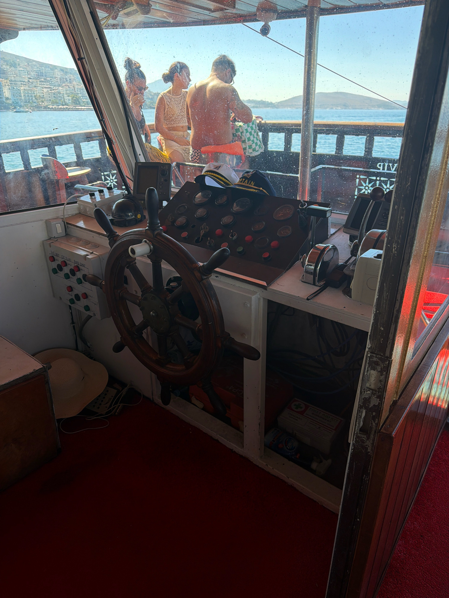 The image shows the interior of a boat's bridge, likely in the city of Saranda, Albania, given the clear view of the coastline with buildings on a hillside and the Mediterranean Sea. The bridge is equipped with a wooden steering wheel, a dashboard with various gauges and switches, and a throttle control. In the background, through the windows, three people are visible on the deck of the boat. One person, a shirtless male, is standing and looking towards the sea. Another person, a female, is sitting and looking at the male. A third person, partially obscured, is also visible. A straw hat lies on the floor next to a control panel with red and green buttons. A first aid kit is stored below the dashboard. The overall lighting suggests a sunny day.