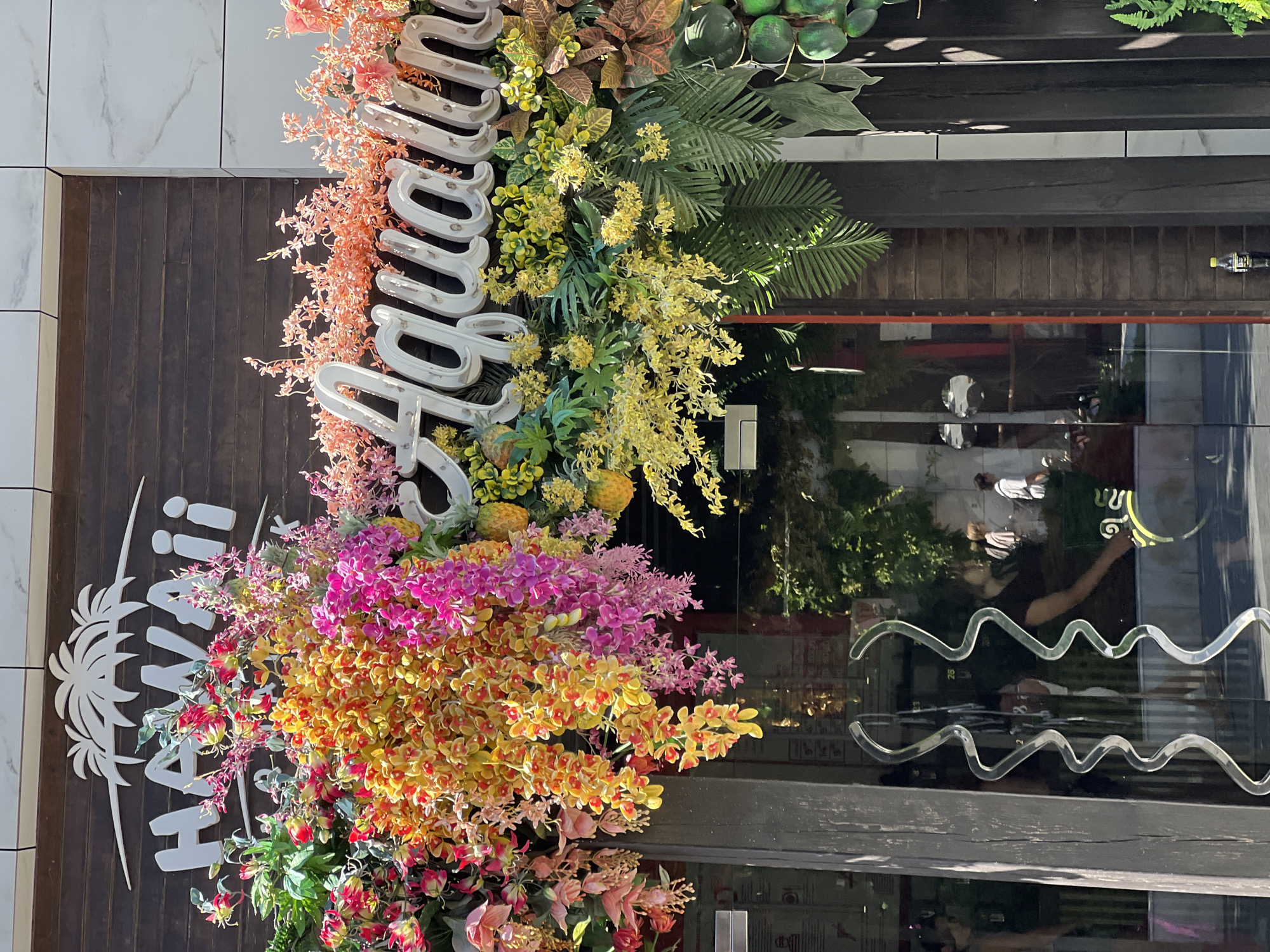 The image shows the facade of a store with a prominent "HAWAII" sign above a decorative floral arrangement and a cursive "Aquamarine" sign. The store entrance is made of glass with ornate, wavy handles. The building's facade features dark wooden planks and white marble tiles. The floral arrangement is abundant with colorful artificial flowers in shades of pink, yellow, and orange, along with green foliage. Reflections in the glass doors show people and the street outside. This establishment is located on Derybasivska Street in Odesa, Ukraine.