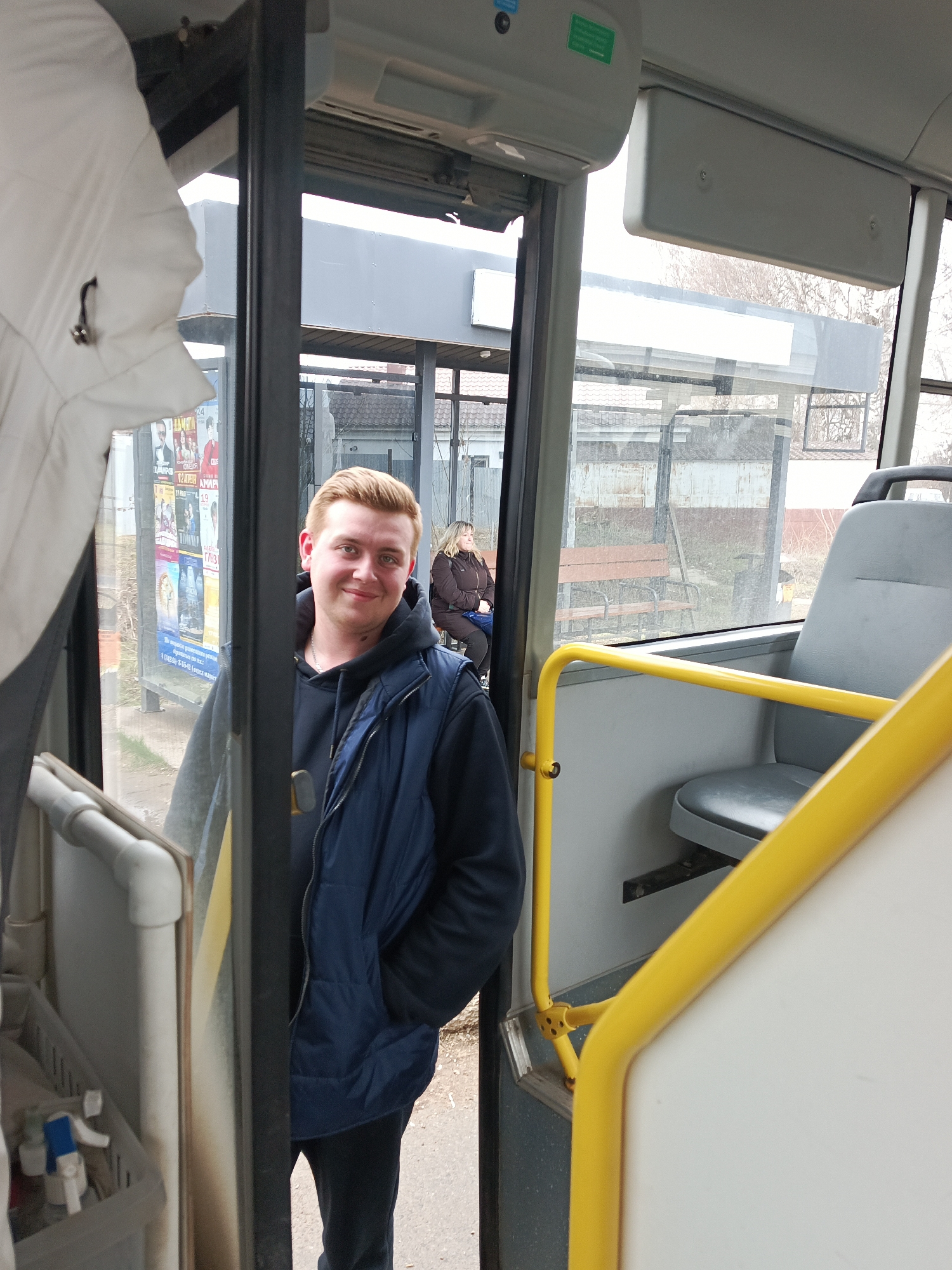 A young man with light reddish-blonde hair and a friendly smile stands in the open doorway of a public bus, looking directly at the camera. He is dressed in a dark blue puffer vest over a grey hooded sweatshirt and dark trousers, with a silver chain visible around his neck and a small tattoo on his left neck. To his immediate left, a white jacket hangs inside the bus.
The bus interior features prominent yellow grab rails, grey seats, and grey overhead panels. Through the open door and large windows, a bus stop is visible. A woman in a brown jacket sits on a wooden bench beneath a bus shelter constructed with metal frames and glass panels. Beyond the bus stop, bare trees and residential buildings with tiled roofs can be seen in the distance, suggesting a semi-urban environment in Chaikovsky, Russia.
Several posters with Cyrillic text and images are affixed to the glass panels of the bus shelter. Readable text includes "24 АПРЕЛЯ" (April 24), "12 МАРТА" (March 12), and "КИНО" (KINO, meaning cinema), indicating advertisements for films or events. The scene is set during the daytime, likely overcast due to the diffused lighting and lack of harsh shadows. The attire of the individuals suggests cool weather, possibly late autumn, winter, or early spring. The bus appears to be stationary at the stop.
