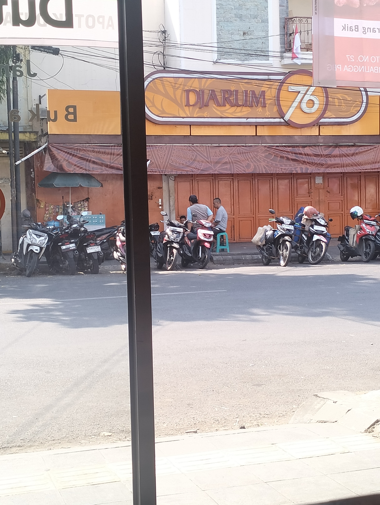 In Purbalingga Kulon, Indonesia, a street scene unfolds in front of a building displaying the prominent "Djarum 76" logo, a brand associated with tobacco products. Parked motorcycles line the sidewalk, suggesting a common mode of transportation and a potentially busy area. Two men are visible near the entrance of the building, seated on a stool and a motorcycle respectively, appearing to be in conversation or resting. The architecture suggests a commercial establishment, possibly a store or office, with a large, roll-up metal door partially visible. The surrounding environment appears to be urban, with a street in the foreground and hints of buildings and wires overhead, indicative of a typical Indonesian city street. The lighting suggests it is daytime, with shadows cast by the motorcycles and the building.