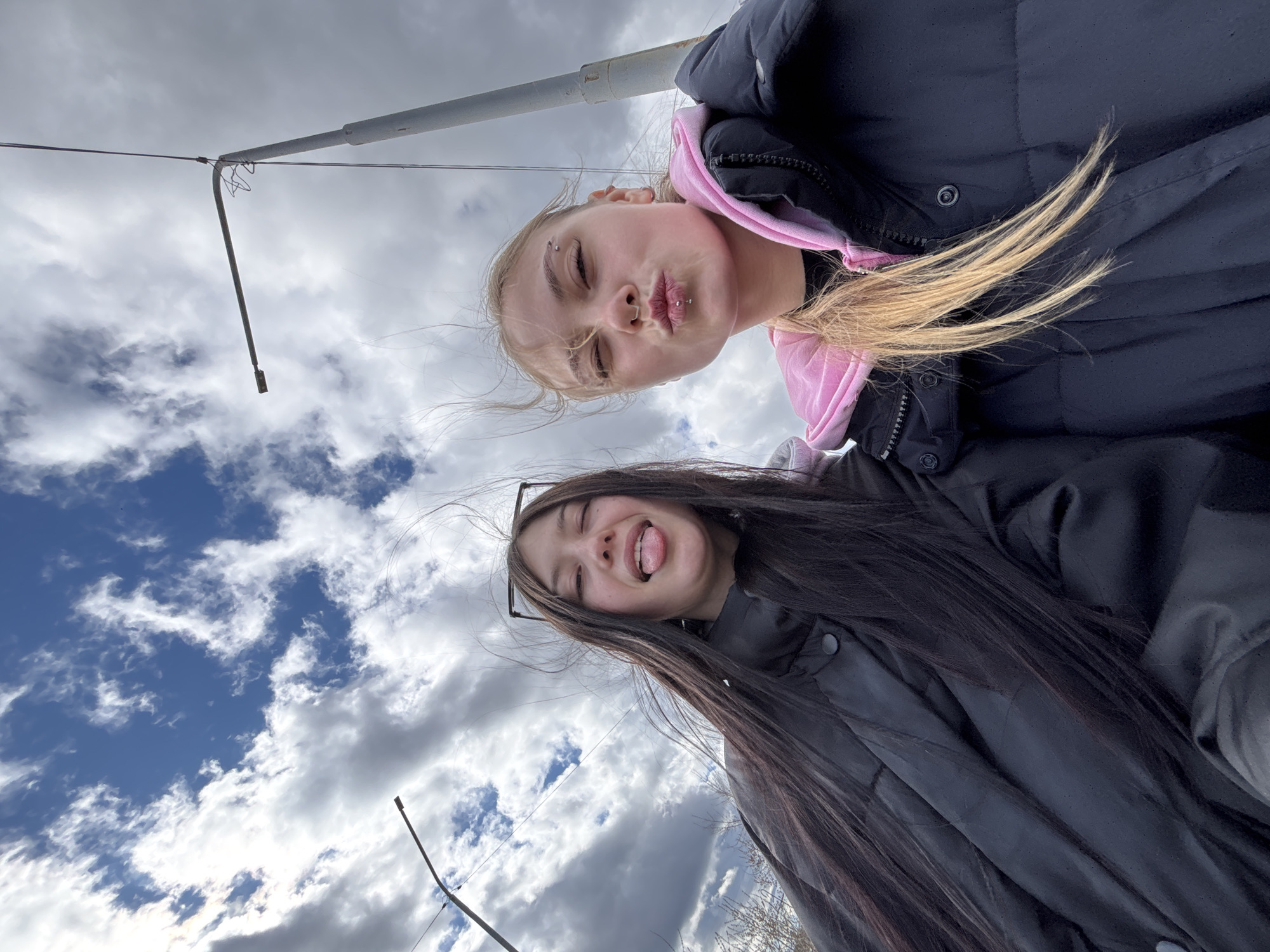 Two young women are positioned close together in an outdoor selfie taken from a low angle. One woman, with fair hair and a pink hood, is puckering her lips. The other, with long dark hair, is sticking out her tongue. They are both wearing dark jackets. The sky is visible behind them with light and dark clouds. There is a streetlight pole in the background. The image was taken in Chaikovsky, Russia.