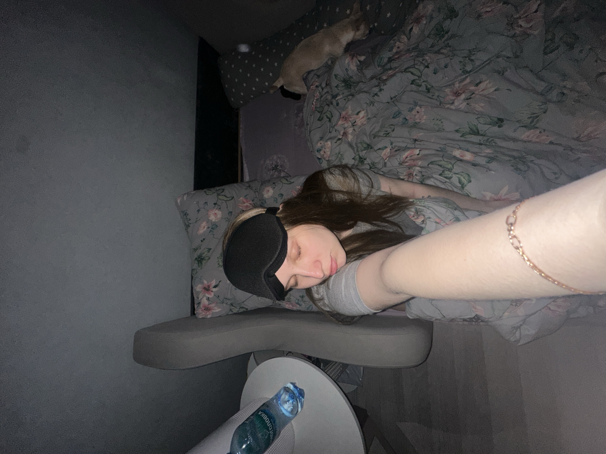 The image, taken in a bedroom in Chaikovsky, Russia, is tilted to show a young woman lying on her side on a bed, appearing to be asleep or resting. She wears a black sleep mask over her eyes and a grey t-shirt. Her long, dark brown hair is spread around her head. Her left arm extends towards the foreground, displaying a delicate gold-colored bracelet on her wrist.
The bed features a grey duvet cover and a pillow with a floral pattern of pink and green flowers. Behind the woman, a small, light-colored cat is resting on the bed, near another pillow with a grey and white star pattern. Adjacent to the bed, a round, light-colored table or stool holds a plastic water bottle with a blue cap. Partially visible Cyrillic text on the bottle reads "БОЛОЖСКАЯ" (Bolozhskaya). The room's walls are a dark grey. The dim lighting, the sleep mask, and the presence of the sleeping cat suggest it is nighttime or a very subdued, quiet environment. The overall scene conveys a sense of peaceful rest.