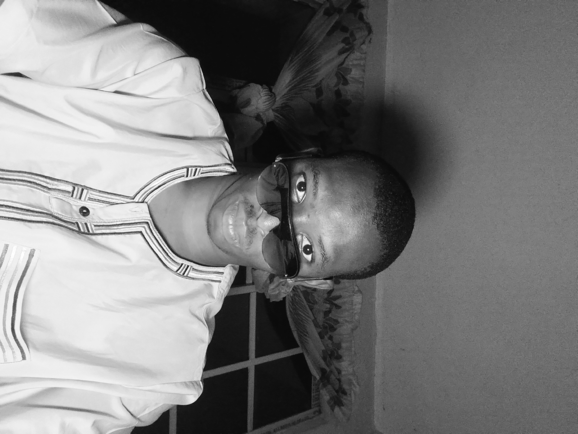 A black and white, head-on, medium shot photograph shows a young Black man wearing sunglasses and a light-colored tunic with decorative trim around the neckline and pocket. He is looking directly at the camera, with his mouth slightly open. The background consists of a window with a patterned curtain. The image appears to have been taken indoors, possibly in Lagos, Nigeria.