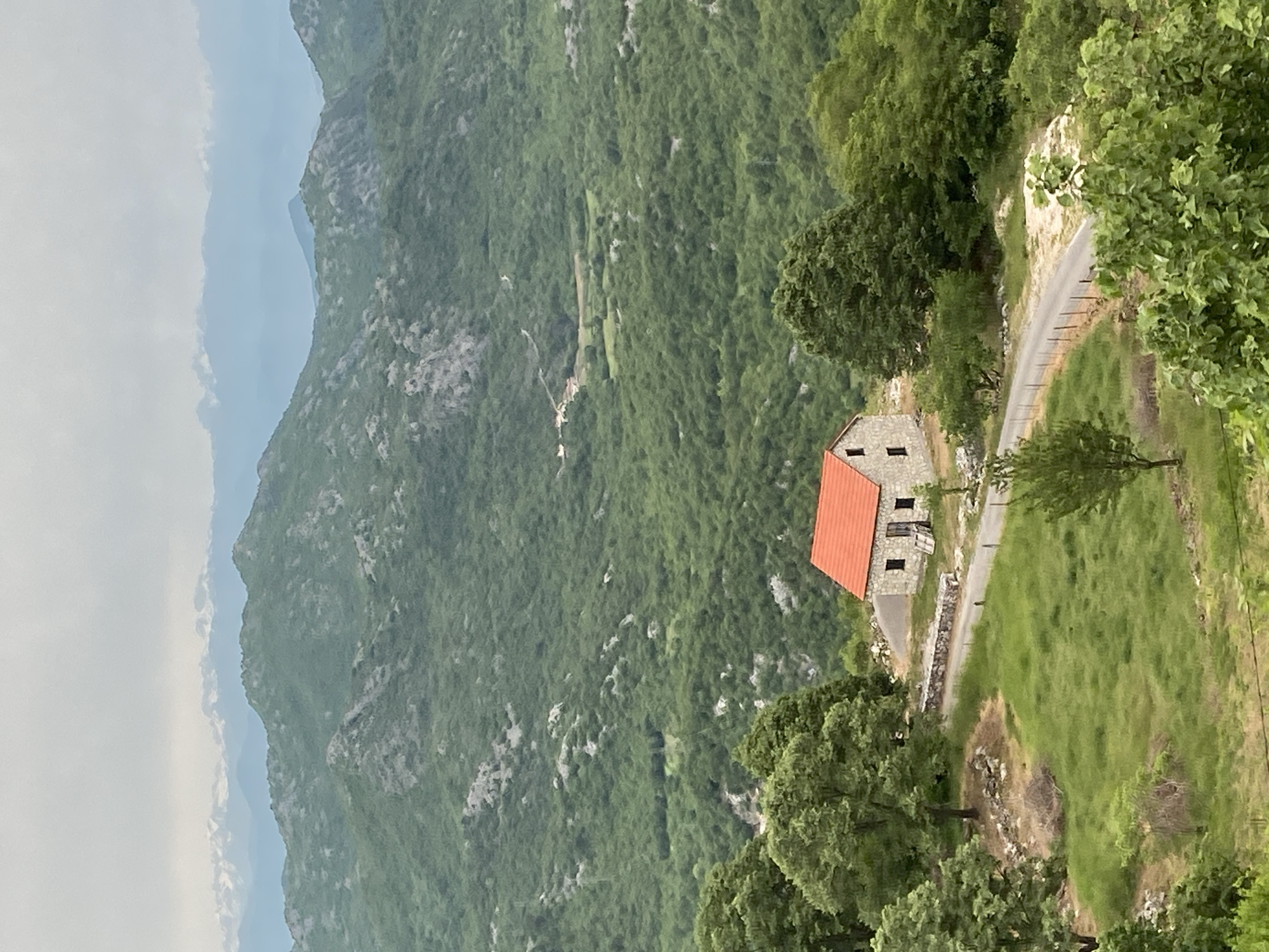 An elevated, outdoor, medium-shot depicts a stone house with a red tiled roof, located in Lipa Dobrska, Montenegro, set amidst a steep, green, mountainous landscape. A narrow, winding road passes by the house, flanked by grassy terrain and scattered trees. In the background, layers of mountains fade into the hazy sky.