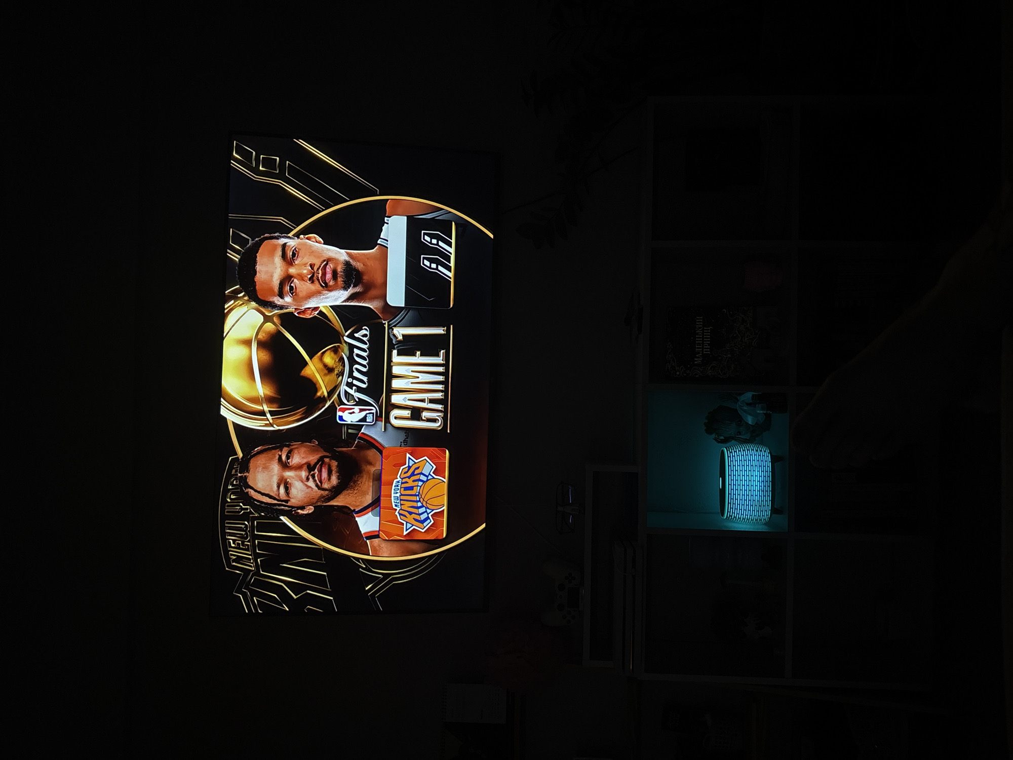 A dimly lit room in Zheleznogorsk, Russia, features a large television screen on the left, displaying a vibrant basketball graphic, and a white shelving unit on the right. The television screen is rotated 90 degrees counter-clockwise from its intended viewing orientation but shows a graphic for "NBA Finals GAME 1." Two male basketball players are prominently featured: one on the left (bottom-right in the original image orientation) associated with the "NEW YORK KNICKS," and another on the right (top-left in the original image orientation) wearing a jersey with the number "11." The words "Finals" and "GAME 1" are vertically displayed between the players, next to a golden basketball design.
To the right of the television, a white modular shelving unit is visible. Within one of the middle compartments, a cylindrical object emits a bright, blue-green glow. A hand is partially visible, reaching towards this glowing item. In the top-left compartment of the shelving unit, a book with Cyrillic text, "МАЛЕНЬКИЙ ПРИНЦ" (Malenkiy Prints, meaning "The Little Prince"), is discernible, affirming the Russian location context. Other shelves contain various dark, indistinct objects, possibly electronic devices. The room's low light levels, illuminated primarily by the TV screen and the glowing object, suggest a nighttime setting. The scene captures a moment of viewing sports entertainment in a personal space.