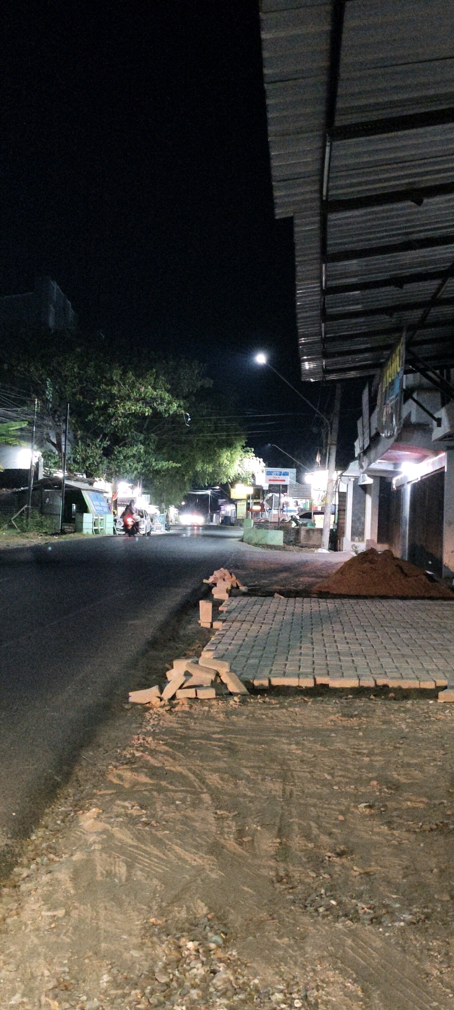 At night in Leteh, Indonesia, a dark asphalt road is visible on the left side of the frame. Along the right side of the road, there is a construction area. Partially laid paving stones form a neat rectangle. To the right of the paving stones, a conical mound of reddish-brown soil is piled high. Scattered bricks lie on the edge of the paved area and on the surrounding rough ground. A row of buildings with corrugated metal roofs lines the right side of the street, with illuminated storefronts suggesting commercial activity. Trees with dense foliage are visible on the left side of the road, illuminated by streetlights. Traffic lights and street lamps cast a faint glow on the street, where a few vehicles, including a motorcycle, are in motion. The overall scene depicts a street in a town, with ongoing construction work alongside existing businesses.