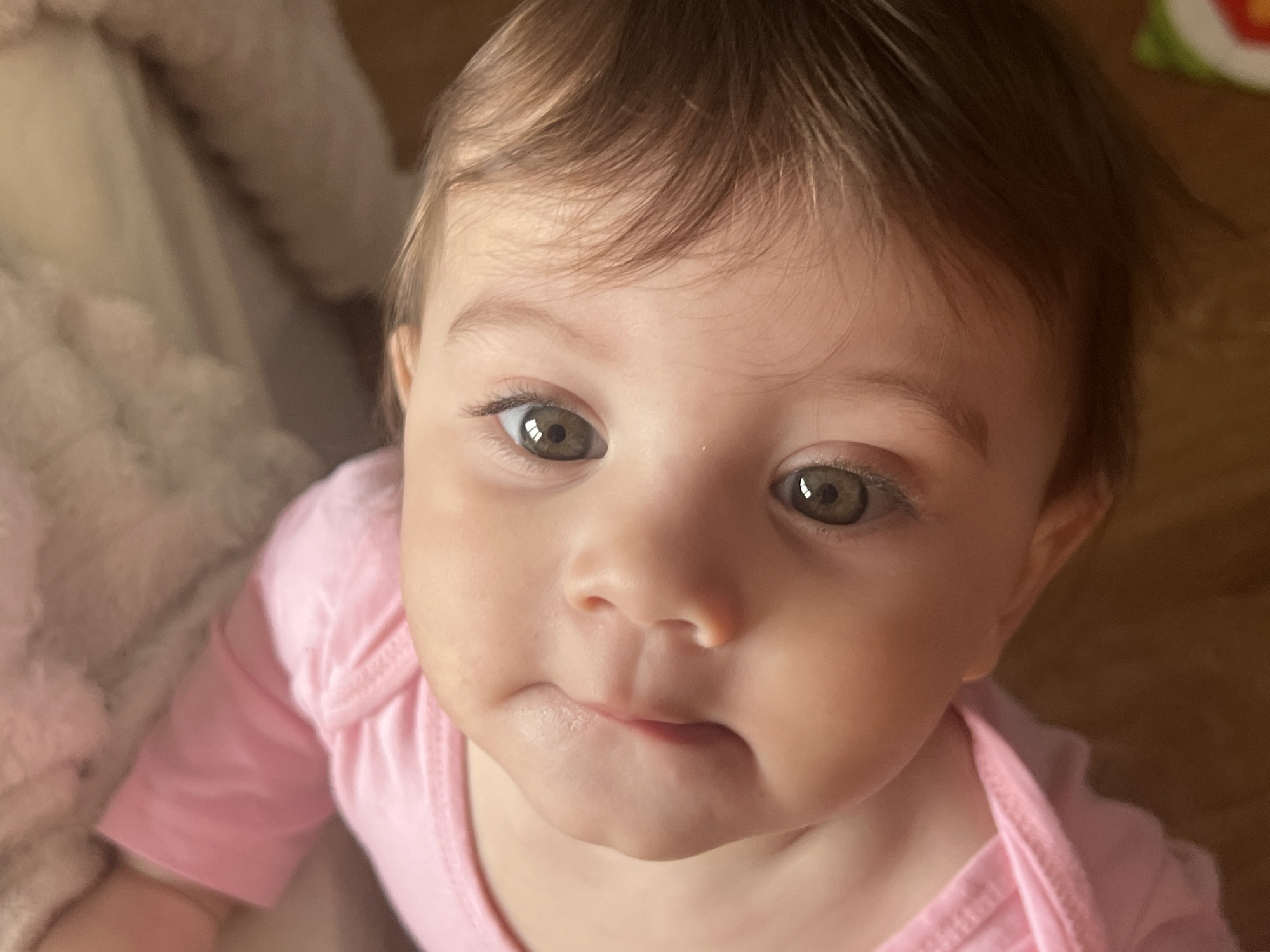 A close-up, high-angle shot focuses on a baby with light brown hair and hazel eyes. The baby is wearing a pink onesie. The background shows a cream-colored fuzzy blanket on the left and a wooden floor on the right.