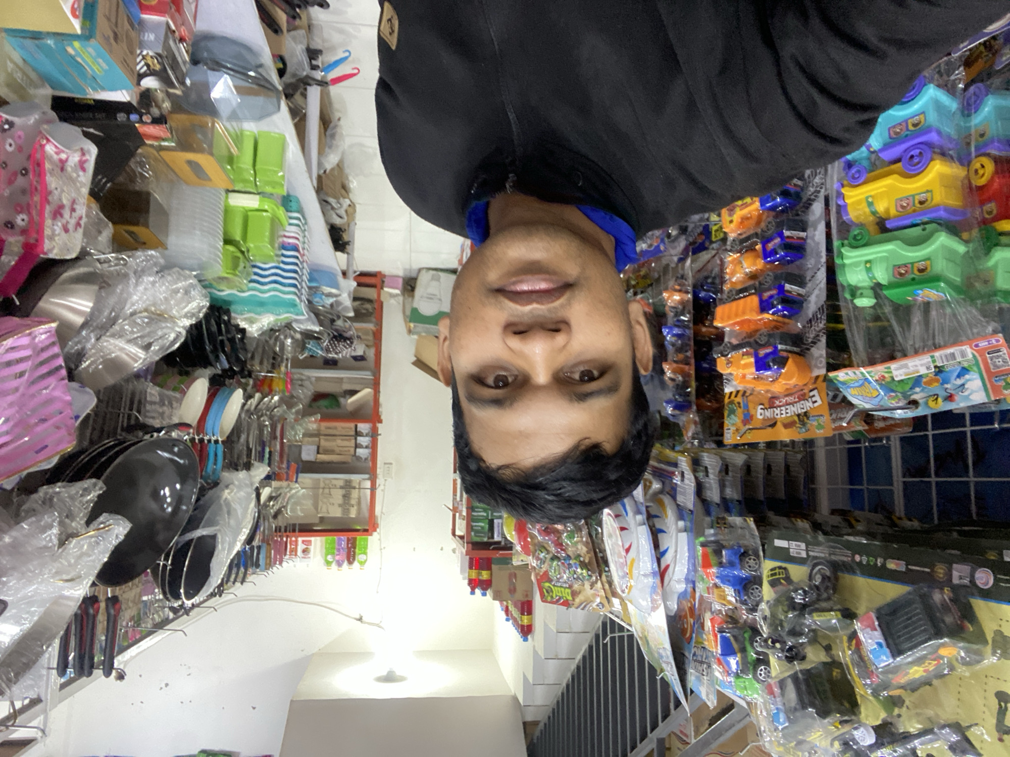 The media file is a selfie taken by a man in a store. The man is wearing a black jacket over a blue shirt and is looking directly at the camera. His face is partially visible due to the image being rotated. The background is filled with shelves displaying various items, including kitchenware, plastic containers, and toys.
In the foreground, on the left side, there are pots and pans wrapped in plastic. To the right of these, there are shelves with kitchen utensils like spatulas and ladles. Further to the right, shelves are stocked with colorful toys, including toy trucks and stacking toys, some of which are packaged in clear plastic. There are also various bottled liquids, possibly cleaning supplies or beverages, on some shelves. The setting appears to be a retail store in Lamteumen Barat, Indonesia. The lighting suggests it could be daytime, though no specific weather details are apparent. The man's expression is neutral. Visible text includes "ENGINEERING TRUCK" on a toy packaging.