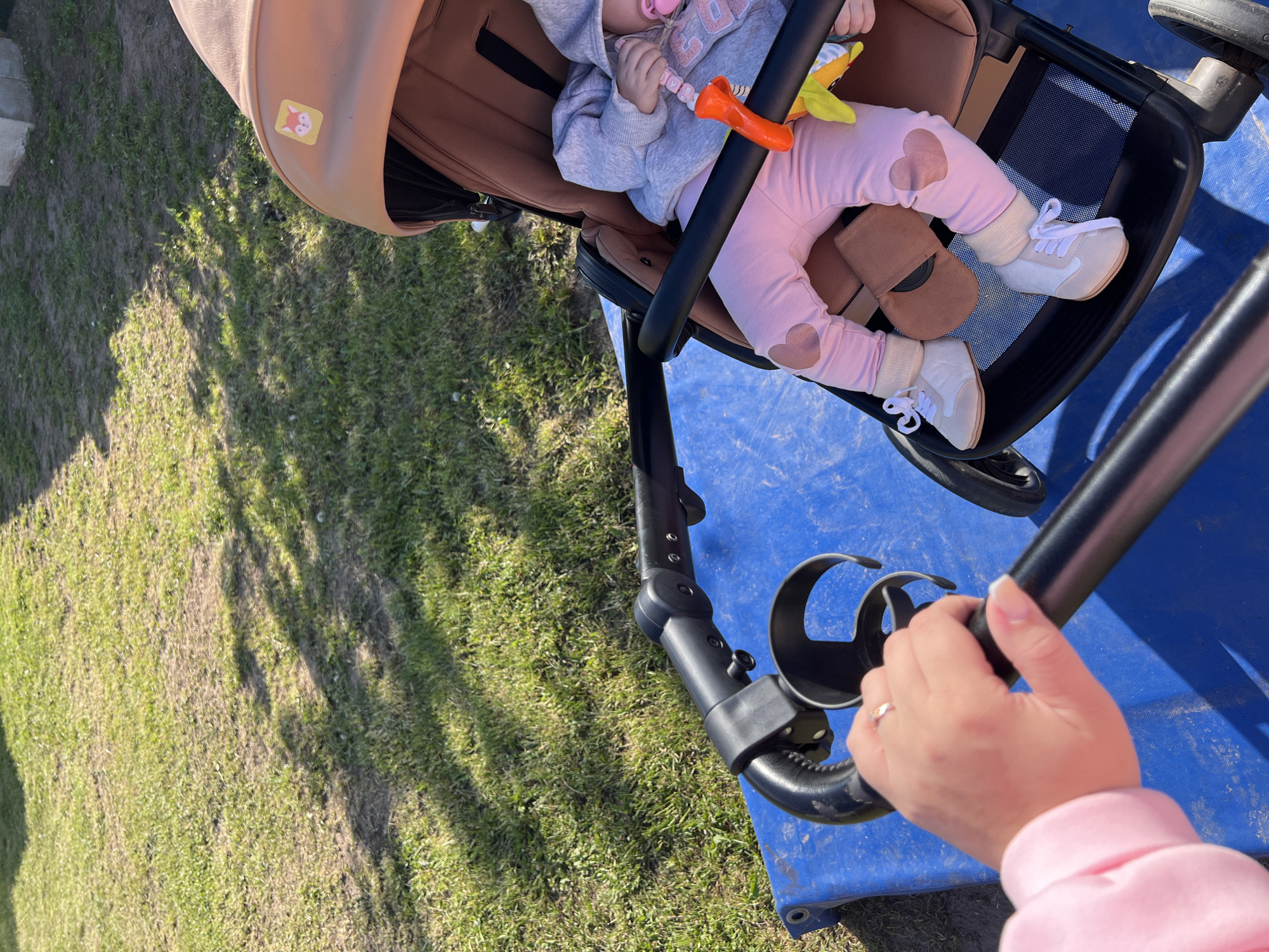 The image shows a baby in a stroller on a blue surface, potentially a playground area. The baby is wearing a gray hooded sweatshirt and pink pants with heart details on the knees. The baby also has on white shoes and is holding an orange toy. The stroller has a brown hood and a black frame. A hand with a ring is holding the handle of the stroller. The background includes a grassy area and the photo was taken in the city of Chaikovsky, Russia.