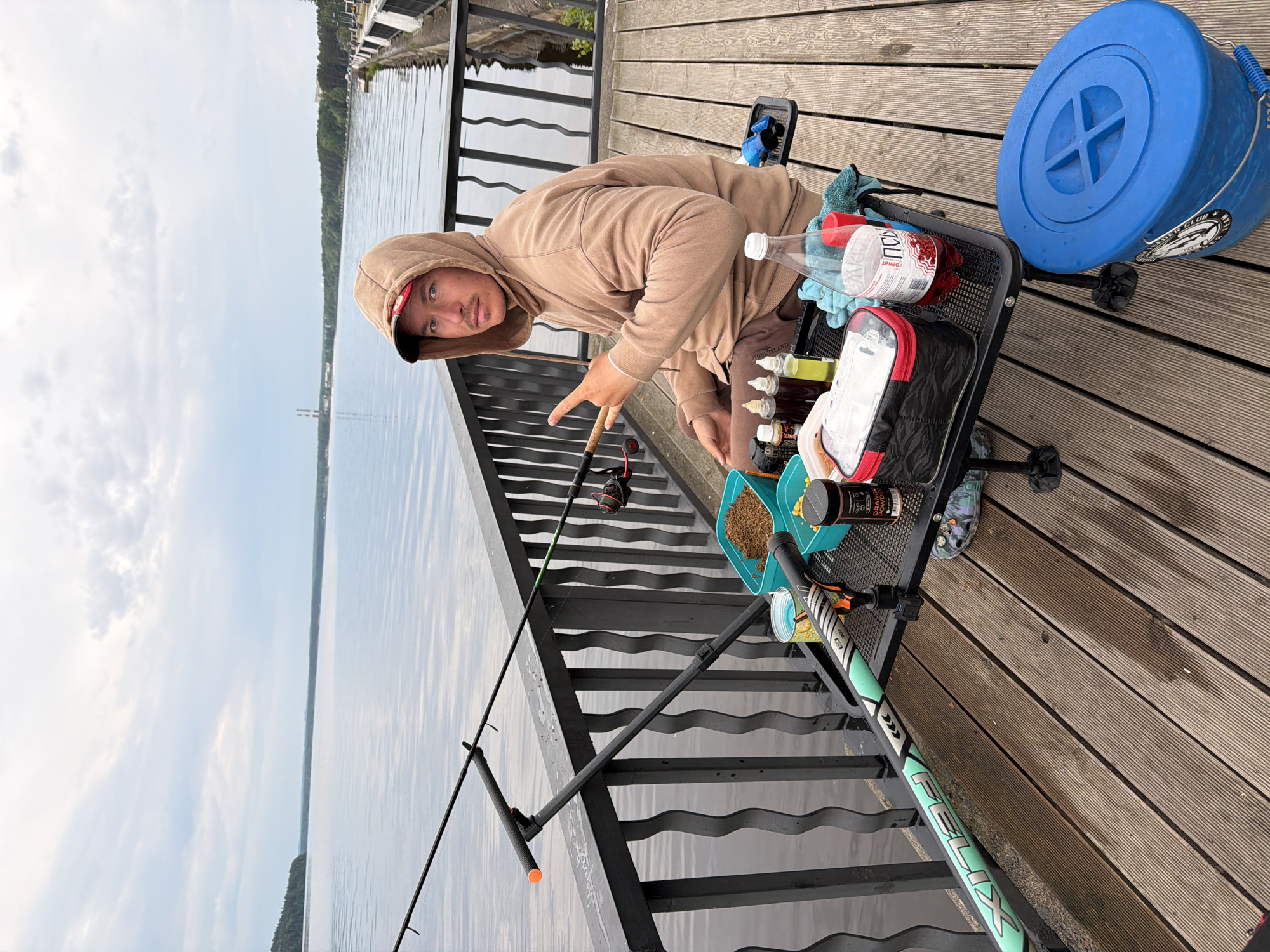 The image captures a young man fishing on a wooden pier or deck by a large body of water in Chaikovsky, Russia. The scene is set outdoors under an overcast sky, with a calm body of water stretching into the distance and a forested shoreline visible on the far bank.
The main subject is a man wearing a beige hooded sweatshirt, dark pants with the "STONE ISLAND" logo visible, and a cap underneath his hood. He is positioned on the right side of the frame, looking directly at the camera with a neutral expression. He holds a fishing rod in his left hand and makes a gesture with two fingers of his right hand.
Beside him is a black utility trolley or cart filled with various fishing equipment. Notable items include a clear plastic bottle containing a red liquid labeled "ГРАНАТ" (Pomegranate) and "СОК" (Juice). There's also a black tackle bag with red accents, several small plastic bottles likely holding fishing attractants, and two light blue containers, one filled with brown ground bait and the other with yellow pellets. A black container with "ORANGE" printed on it is also visible. A large blue plastic bucket with a lid and a partially obscured sticker is on the wooden deck next to the trolley.
Another fishing rod, featuring the brand "FELIX," is set up on a rod stand that extends over the water, parallel to the dark metal railing of the pier. The wooden decking beneath the man and his gear suggests a well-maintained fishing spot. The overall ambiance indicates a quiet, daytime fishing excursion.