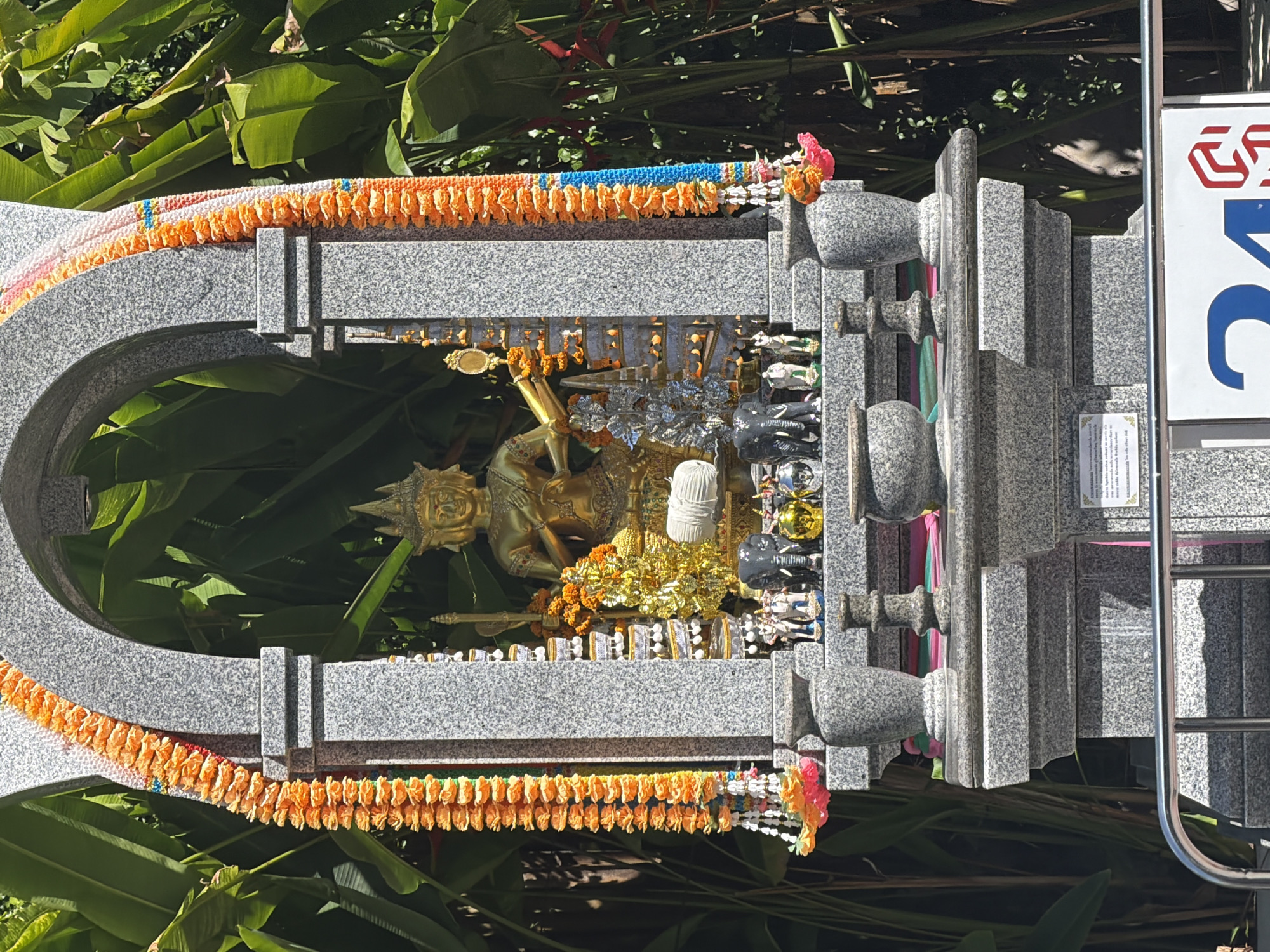 This is a medium shot of an outdoor shrine in Baan Chaweng, Thailand. The shrine is made of grey granite and features an arched opening. Two garlands of orange marigold flowers with blue and white beads hang from the sides of the arch. Inside the shrine, a golden statue of a deity with multiple faces and hands is seated. The deity is adorned with a crown and jewelry. In front of the statue are offerings, including small elephant figures, golden and silver decorative items, and a white cylindrical object. The shrine is situated in front of lush green foliage, likely tropical plants. In the foreground, a silver metal railing is visible, and behind it, a sign with a blue and red logo is partially visible. The lighting suggests it is daytime with strong sunlight.
