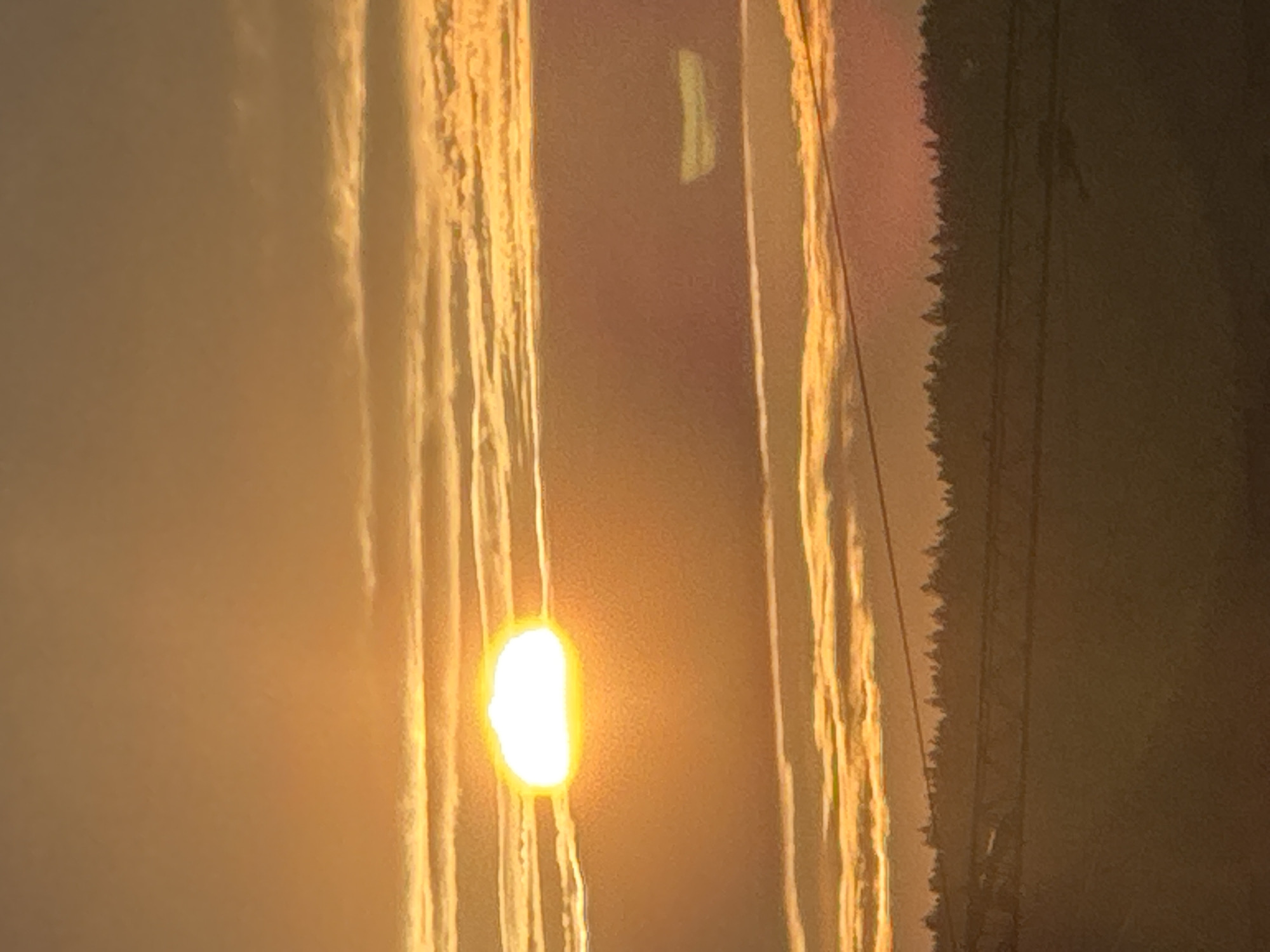This is a photograph of a sunset in Pernik, Bulgaria. The sun is partially obscured by horizontal clouds, creating a warm orange and yellow glow across the sky. There are several layers of clouds, some appearing wispy and others thicker. In the foreground, a dark silhouette of a treeline is visible against the sky, possibly a forest or a row of trees. Below the treeline, there appear to be power lines or some form of aerial infrastructure. The lower portion of the image is darker, suggesting the ground or a landscape feature. The overall impression is one of a natural landscape at dusk.