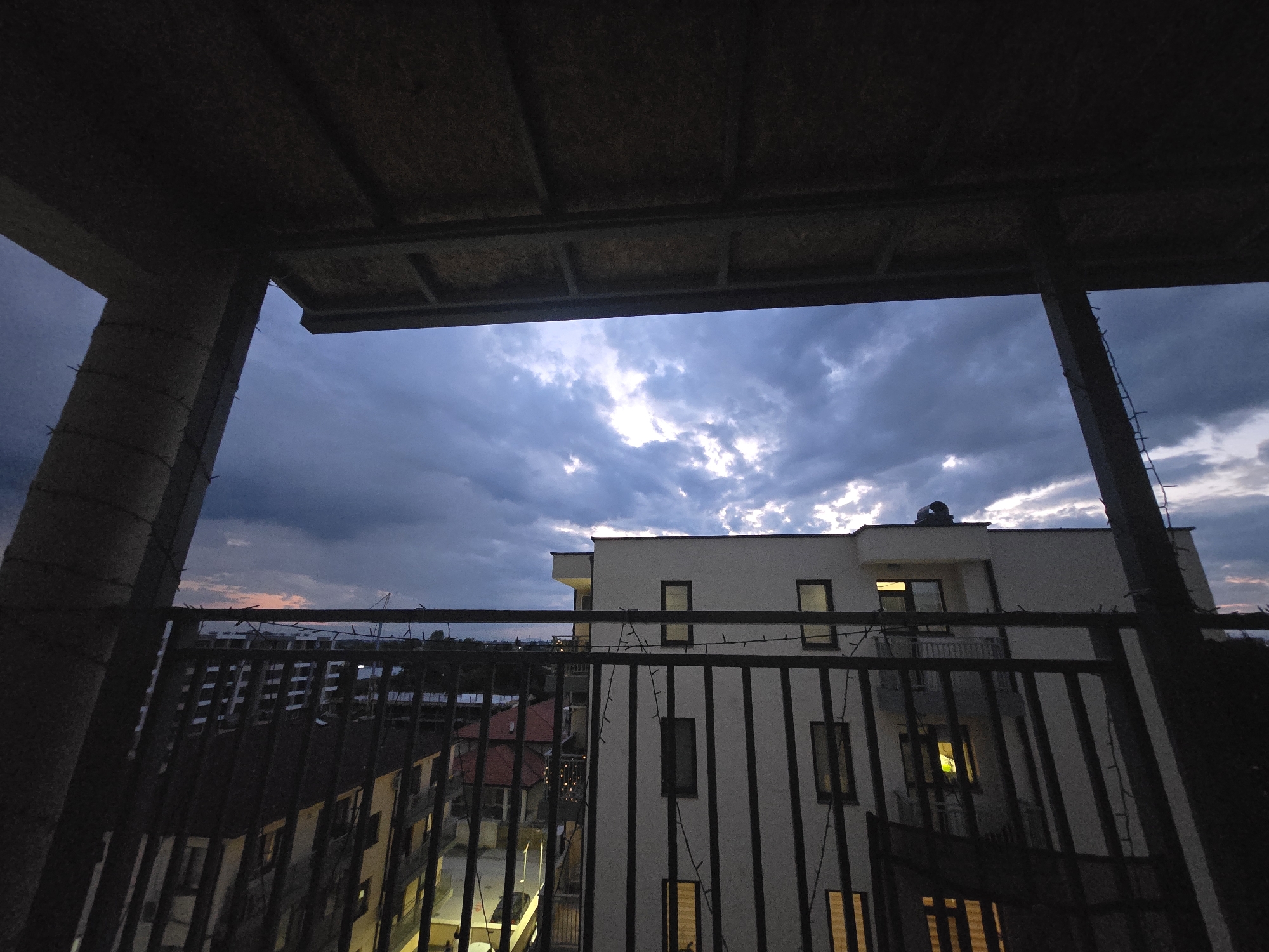 The image captures an urban or suburban landscape at dusk or dawn, viewed from a shadowed balcony. The foreground is dominated by the dark, structural elements of the balcony, including thick concrete pillars on the left and right, and a dark, possibly corrugated ceiling overhead. A black metal railing with vertical bars stretches across the mid-ground, draped with string lights.
Beyond the railing, a dramatic sky fills the upper half of the scene, characterized by heavy, dark blue-gray clouds with intermittent bursts of bright, almost white light breaking through, suggesting a dynamic weather pattern. In the middle ground, a prominent light-colored, multi-story building stands out against the sky. This building features dark-framed rectangular windows, several of which are illuminated with warm light from within. A small exhaust vent or pipe is visible on its rooftop. Below this building, other structures with darker facades and one with a reddish roof are partially visible. Further in the background on the left, a dense cluster of other dark buildings suggests a populated area. In the far distance on the left, a faint reddish-orange glow on the horizon hints at either sunrise or sunset.
No people are visible. The scene conveys a quiet, transitional moment, with the artificial lights of the buildings starting to punctuate the fading natural light. There is no visible text in the image.