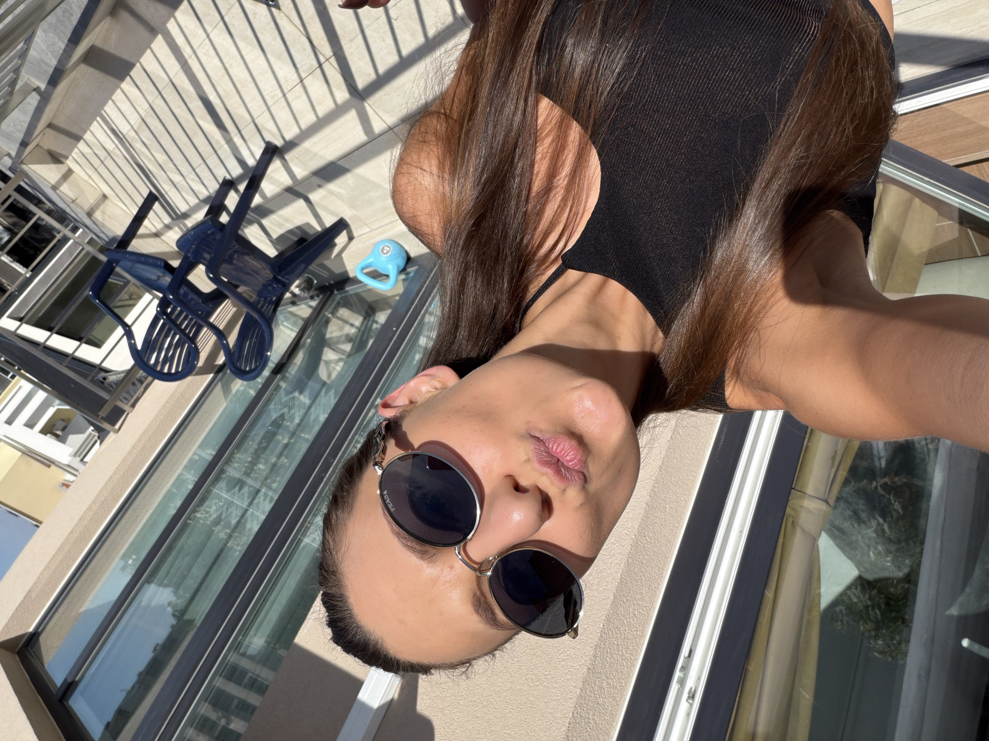 A woman with long, dark hair wearing a black top and sunglasses is taking a selfie on a balcony. She is puckering her lips and looking at the camera. The balcony has light-colored tiled flooring and a metal railing. In the background, there are large glass windows reflecting the sky and surrounding buildings. Two dark blue plastic chairs are visible on the balcony, along with a light blue kettlebell. The scene appears to be in Plovdiv, Bulgaria, given the architectural style of the buildings.