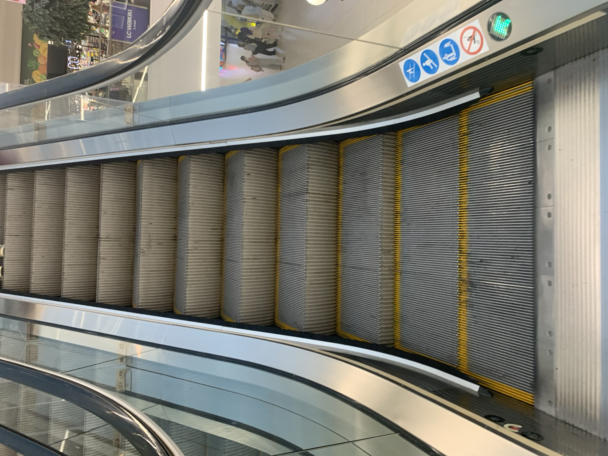 The image shows a close-up view of an escalator in a modern shopping mall, likely in Hrodna, Belarus, given the context of the city mentioned. The escalator steps are made of metallic material with a ribbed texture, and prominent yellow safety lines are visible on the edges of the steps. The escalator is flanked by glass railings and metallic supports, reflecting the surrounding environment. On the right side of the escalator, near the bottom, there are several circular blue and white safety signs, including one with a prohibition symbol indicating no running or carrying items that could obstruct others. A small green light is also visible on the railing. In the background, through the glass, people can be seen walking in the mall, and a sign for "LC Waikiki" is partially visible. The lighting appears to be artificial, typical of an indoor commercial space.