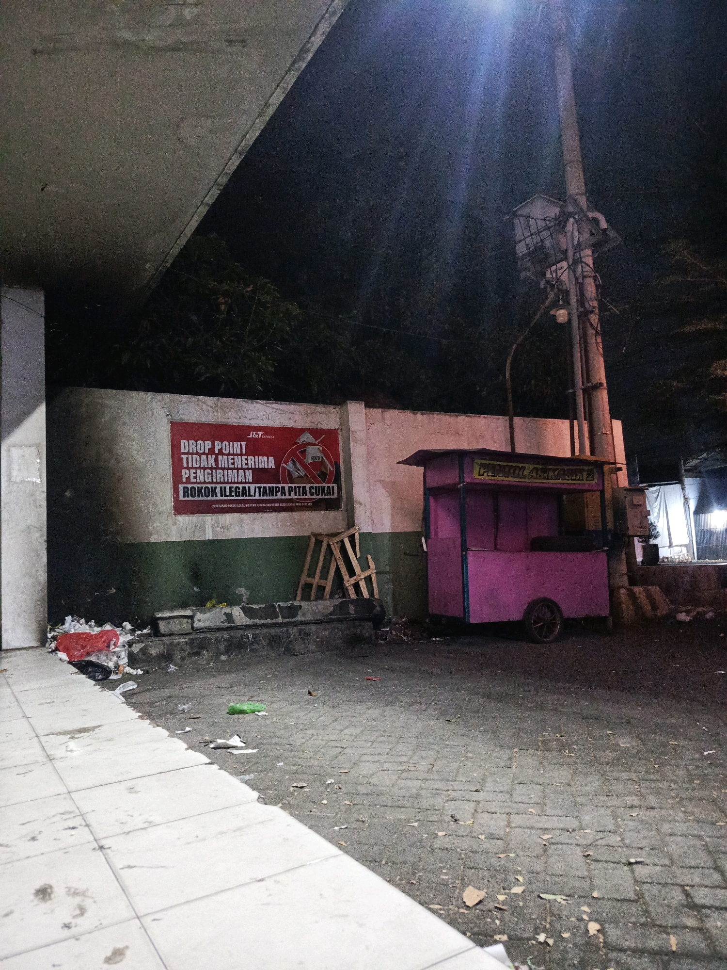 The image depicts a nighttime scene on a street in Pamekasan, Indonesia. A large, red banner is mounted on a wall, bearing the J&T Express logo and text in Indonesian stating that the drop point does not accept the delivery of illegal cigarettes without tax stamps. In front of the wall, there is a pile of trash, including red and black plastic bags, and a small wooden structure. To the right, a pink food cart with the label "PENJUAL ASIKASIK 2" is parked on the paved ground. A tall utility pole with lighting fixtures and electrical equipment stands nearby. The ground is paved with interlocking concrete blocks and is littered with debris, including leaves and paper. The overall environment appears somewhat neglected and dimly lit by a streetlamp.