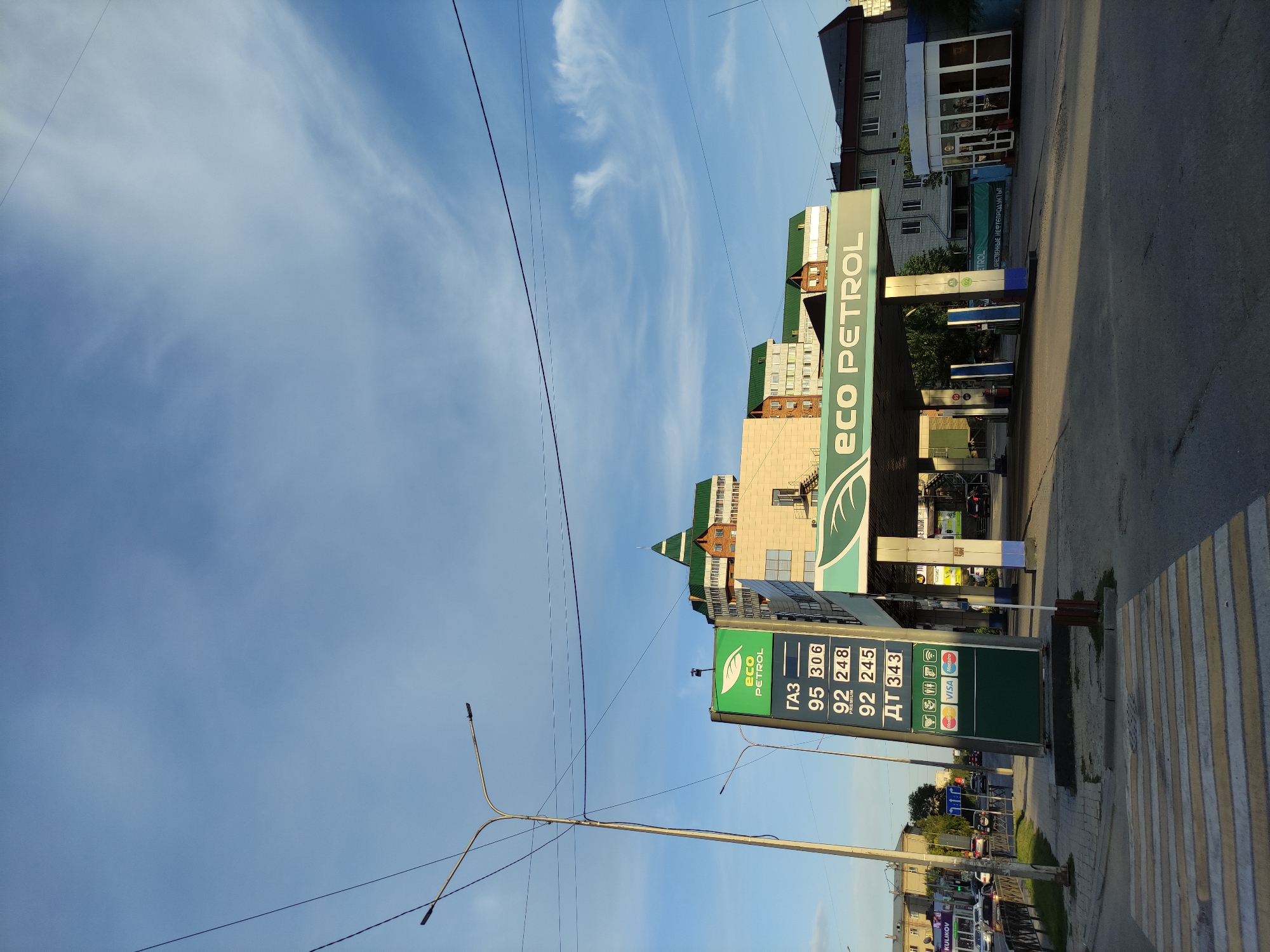 The image captures a daytime scene at an "Eco Petrol" gas station in Semey, Kazakhstan. The sky is predominantly blue with some wispy white clouds. A prominent sign displays fuel prices: GAZ 95 at 306, 92 Premium at 248, and DT at 343. Below the prices, icons for various payment methods, including Mastercard, Visa, and Maestro, are visible.
The gas station canopy features the "Eco Petrol" logo with a green leaf design. Several fuel pumps are positioned under the canopy. In the background, a multi-story building with a distinctive green, peaked roof stands prominently. Adjacent to this building, other structures with light-colored facades and windows can be seen. A street with cars driving and parked is visible to the left, and a pedestrian crossing is marked on the asphalt in the foreground. Streetlights and power lines are present in the frame, indicating an urban environment. The overall scene is of a typical operational gas station.