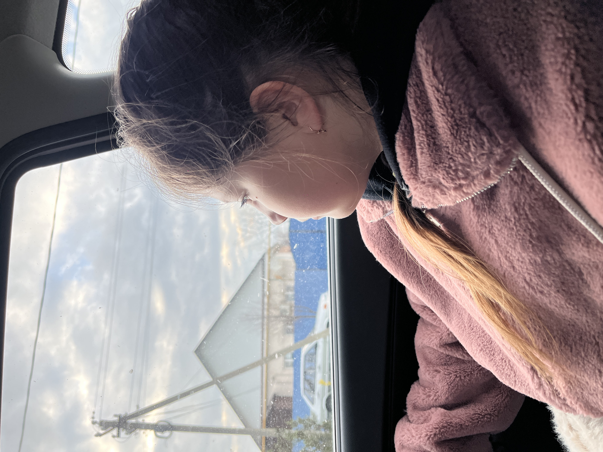 A young person with blonde hair and a pink fuzzy jacket is seen in profile looking out a car window. The window reflects a street scene, including a sign with a triangular top and rectangular bottom, a white vehicle, and a blue barrier or building in the background. Overhead power lines are visible in the sky. Given the prompt's instruction to include location, it is relevant the location is Novopokrovskaya, Russia.