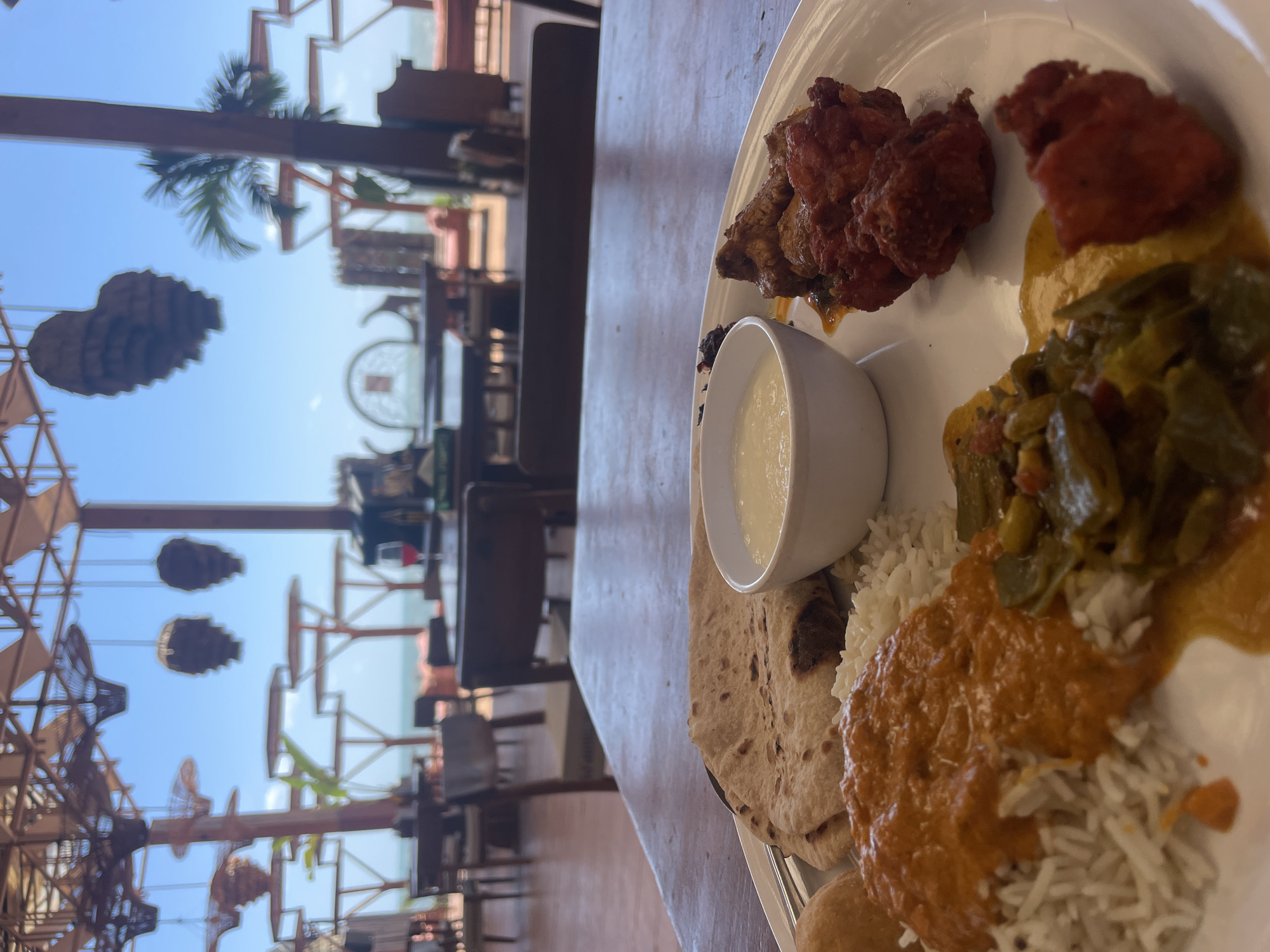 The image shows a partially visible white plate of Indian food on a light wooden table. The food on the plate includes: fried chicken, a red curry dish, a green vegetable dish, rice, roti, and a small white bowl containing a light-colored sauce, possibly yogurt or raita.
The background shows an outdoor seating area, possibly a restaurant or cafe, in Chopdem, India. The outdoor area is decorated with hanging decorative elements, palm trees, and other architectural features. The sky is visible in the background, suggesting a sunny day. Chairs and tables are visible in the out-of-focus background.