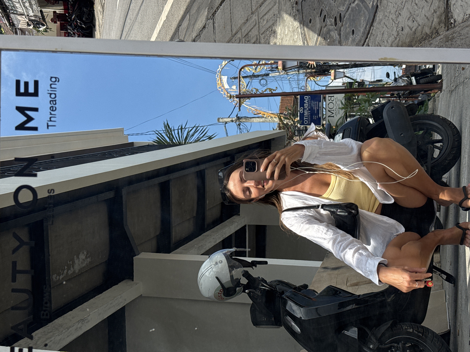 A woman is crouching in front of a black scooter, taking a selfie in a mirror. The mirror reflects a street scene in North Kuta, Indonesia, with a blue sky, power lines, and buildings. The woman is wearing a yellow tank top and a white open shirt, with black shorts and black flip-flops. She is holding her phone in her right hand and wearing earphones. The scooter has a white helmet on the handlebars. A sign in the mirror reads "BEAUTY ON ME" and "Threading".