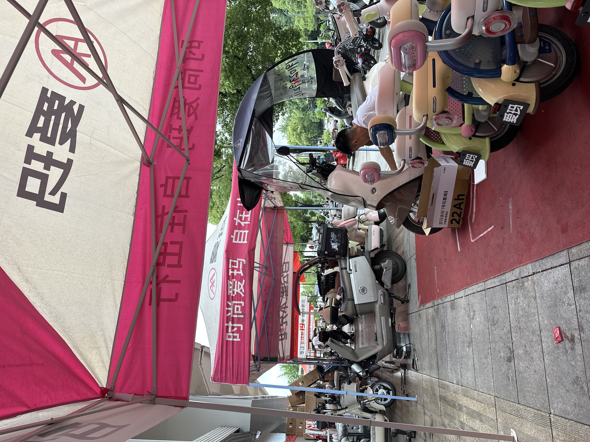The image shows a street scene in Chongqing High-tech Zone, China. Several electric scooters are lined up on a red mat. A person is working on one of the scooters, which has a cardboard box next to it labeled "22Ah." Marketing tents with Chinese writing, including "时尚爱玛 自在" and "爱玛," are set up along the street. Other scooters and pedestrians are visible in the background. The area appears to be a commercial or retail space for electric scooters.