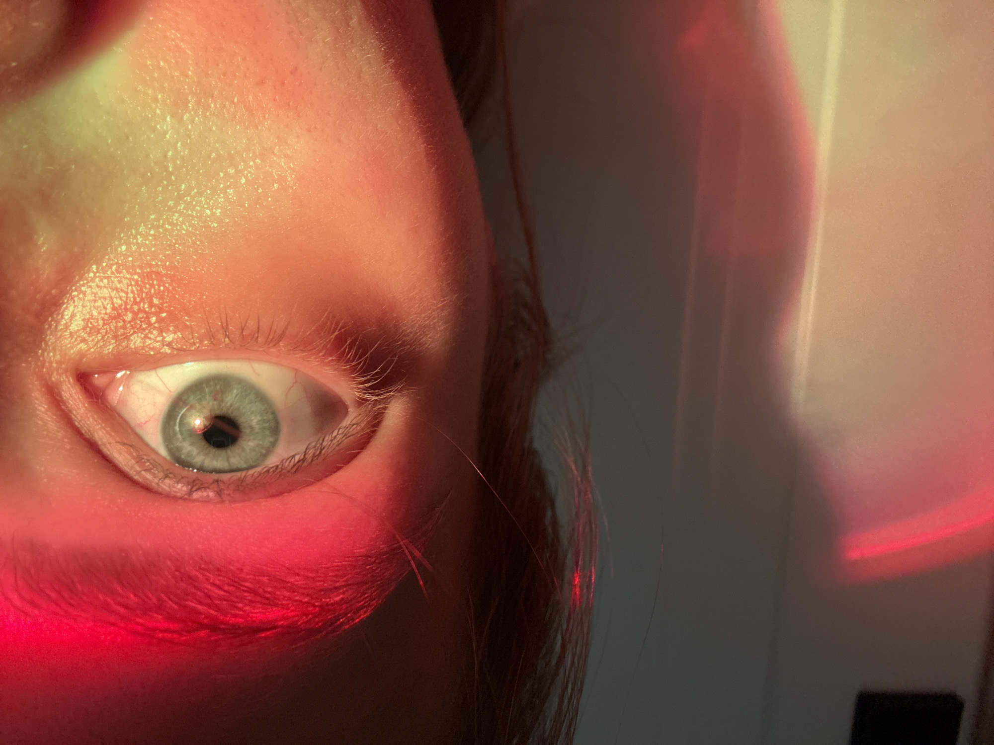 A close-up, vertically oriented photograph captures the left eye and surrounding facial features of a person, illuminated by vibrant, colored artificial light. The eye, featuring a light blue or grey iris and a dark, central pupil, is directed towards the viewer or a light source. Fine red blood vessels are discernible in the white sclera, particularly towards the inner corner of the eye.
The upper part of the face, including the forehead and the skin above the eye, is bathed in a bright, yellowish-green light, highlighting skin texture and faint peach fuzz. Conversely, a striking red light strongly illuminates the lower eyelid, the skin below the eye, and the upper cheek area. This intense red light also extends onto strands of hair visible to the right of the eye.
The background to the right is out of focus, appearing as a blurred expanse of varied light and shadow. Distinct vertical streaks and amorphous patches of red and yellowish light are visible in this background, suggesting reflections off a nearby surface or the spill of the colored light sources. The overall impression is an artistic or experimental portrait, emphasizing the interplay of light, color, and human anatomy. No discernible text is present in the image.