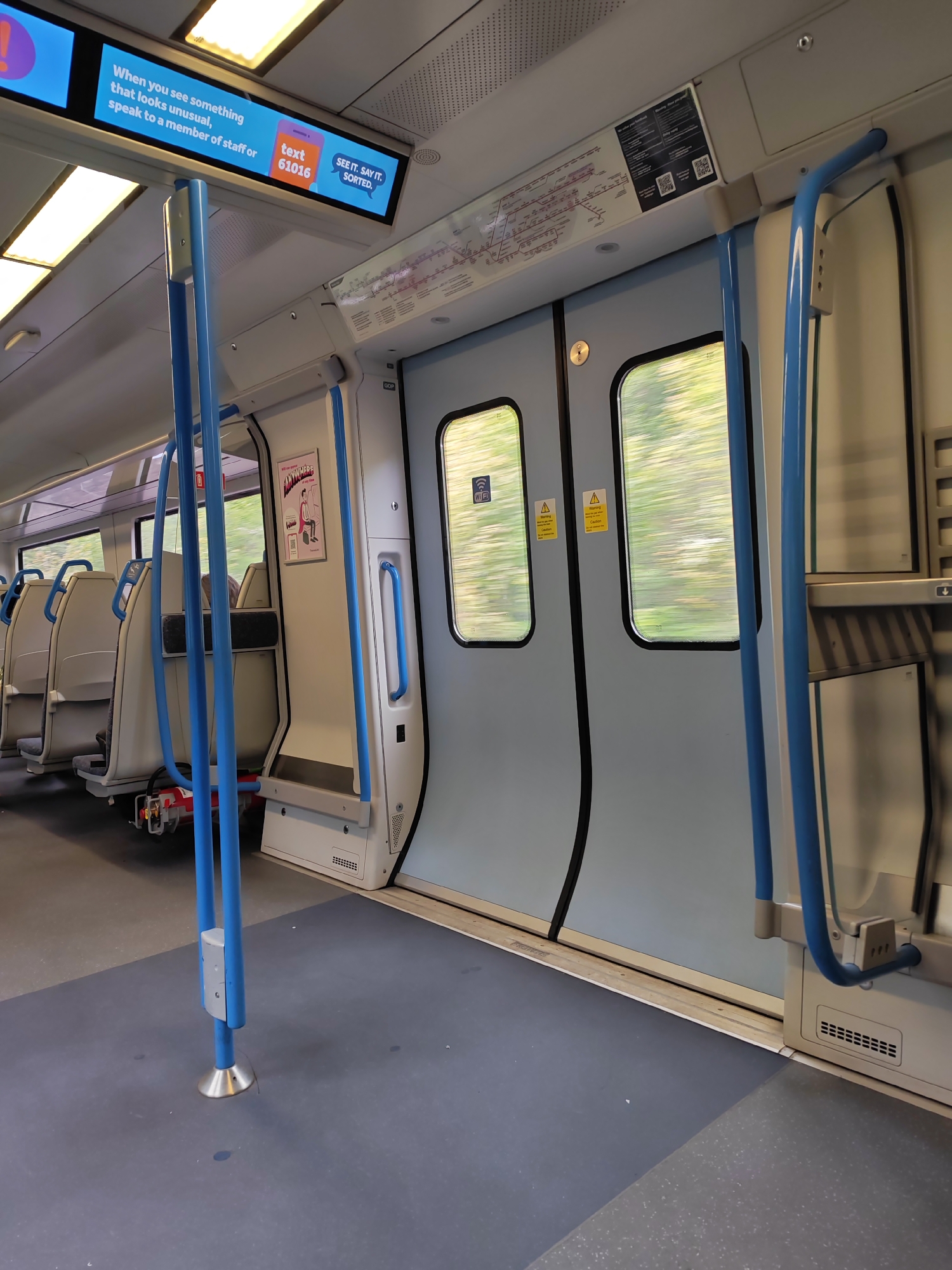 The image captures the interior of a moving train, likely in the borough of Reigate and Banstead, United Kingdom. The train car is mostly empty with unoccupied light-beige seats visible to the left. A vertical blue pole stands in the foreground, firmly secured to the dark-grey floor. Further back, a set of closed sliding doors, painted in a light-grey-blue hue, dominates the scene. Warning signs are visible on the doors. The windows adjacent to the doors show motion blur of green scenery rushing past. On the train's ceiling, illuminated LED displays broadcast a public service message, which reads: "When you see something that looks unusual, speak to a member of staff or text 61016. SEE IT. SAY IT. SORTED." Above the doors are other signs, including a train route map. The carriage appears well-lit, with fluorescent lighting installed in the ceiling.
