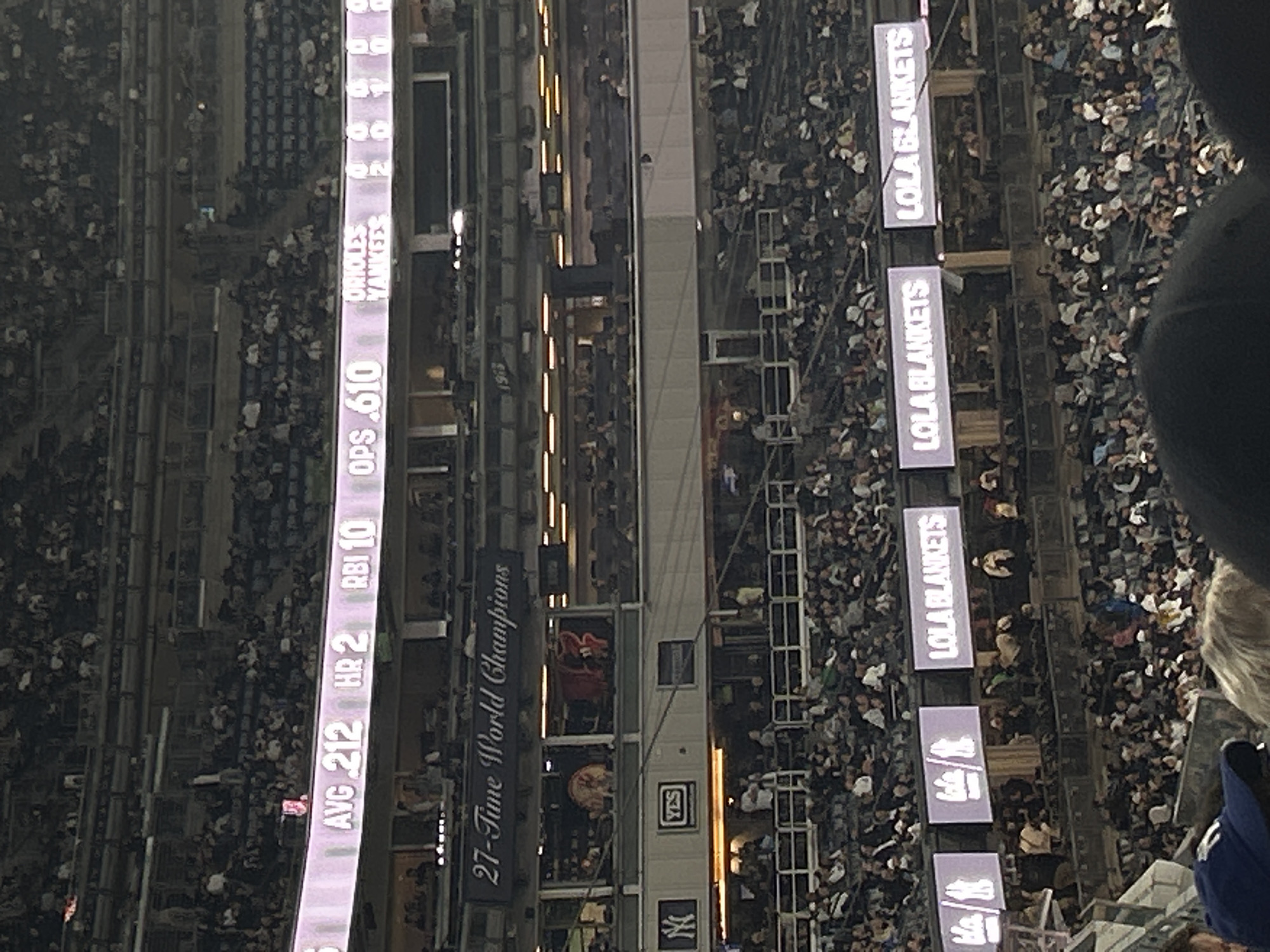 The image is a high-angle shot taken at Yankee Stadium in New York City, depicting a large crowd of spectators. Along the stadium's interior, digital displays present baseball statistics and advertisements. The statistics show the average batting average (AVG) as 212, home runs (HR) as 2, runs batted in (RBI) as 10, and on-base plus slugging (OPS) as .610. There is a banner celebrating the New York Yankees' status as "27-Time World Champions". A "LOLA BLANKETS" advertisement repeats on several digital panels. The Yankees logo is also visible.