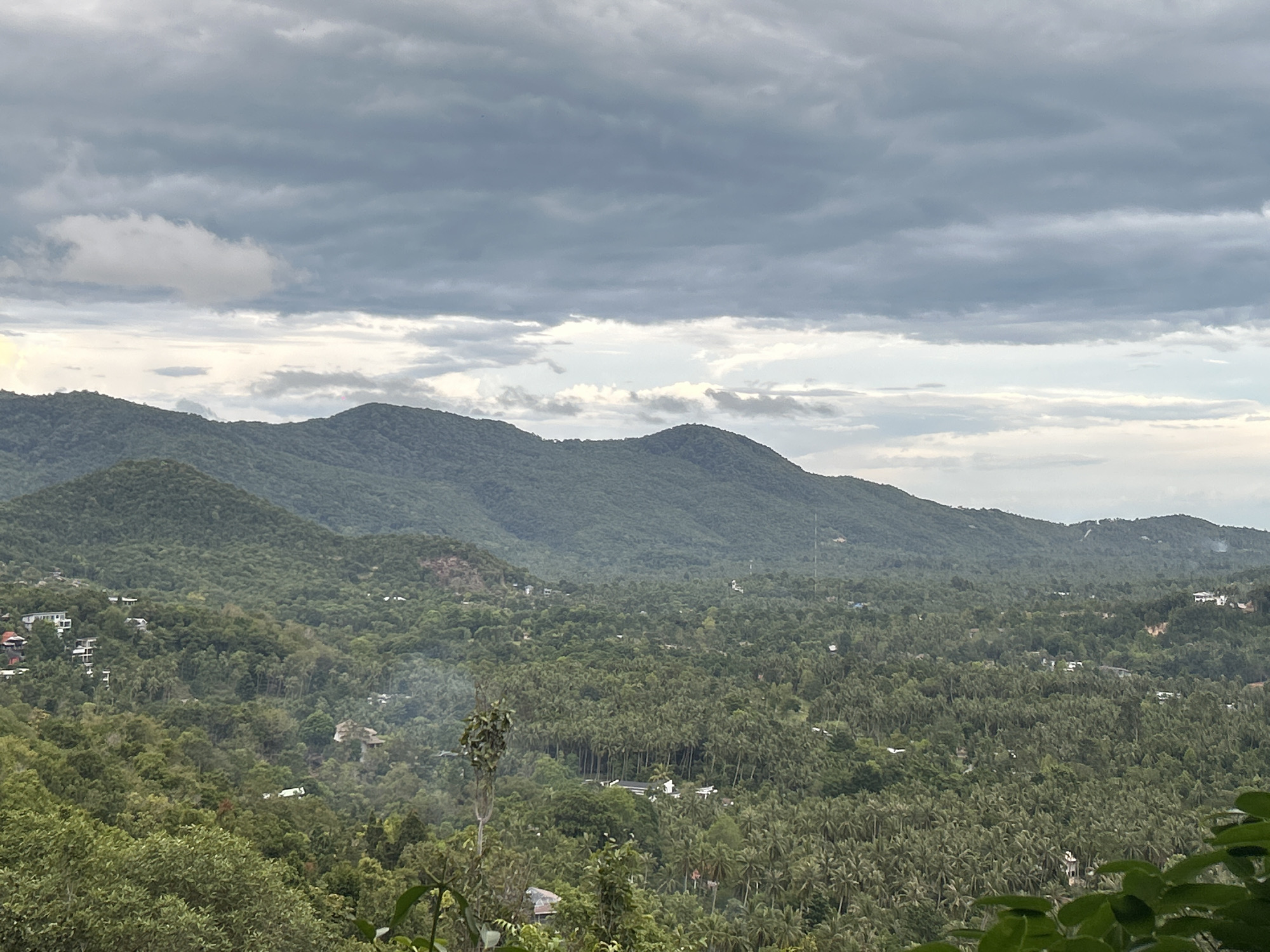 A panoramic view in Baan Hin Kong, Thailand, showcases a densely forested landscape under an overcast sky. Rolling hills covered in lush greenery dominate the middle ground, with individual trees discernible in the foreground and morphing into a solid expanse in the distance. Small structures, likely houses, are scattered sporadically amidst the trees. The sky is heavily clouded with varying shades of gray.
