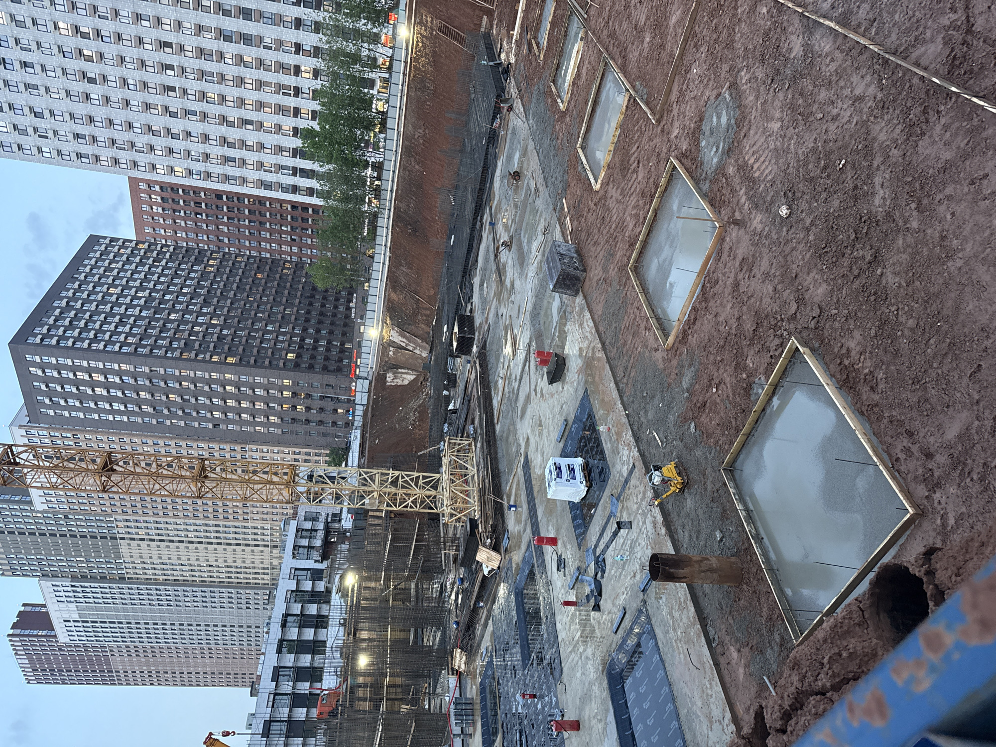 Here's a description of the image:
**Scene:** A construction site in Moscow, Russia.
**Environment:** The site is a large, excavated area with exposed dirt and partially completed concrete structures. Tall apartment buildings surround the site.
**Objects:**
* Several high-rise apartment buildings with repetitive window patterns.
* A yellow construction crane.
* Wooden frames for concrete foundation forms. Some of these are filled with wet concrete.
* Construction materials (possibly insulation) wrapped in white plastic.
* Steel rebar structures, presumably for reinforcing concrete.
* Various construction tools and equipment, including what appears to be a soil compactor.
**Actions:** Construction work is underway, including pouring concrete and setting up structural supports.