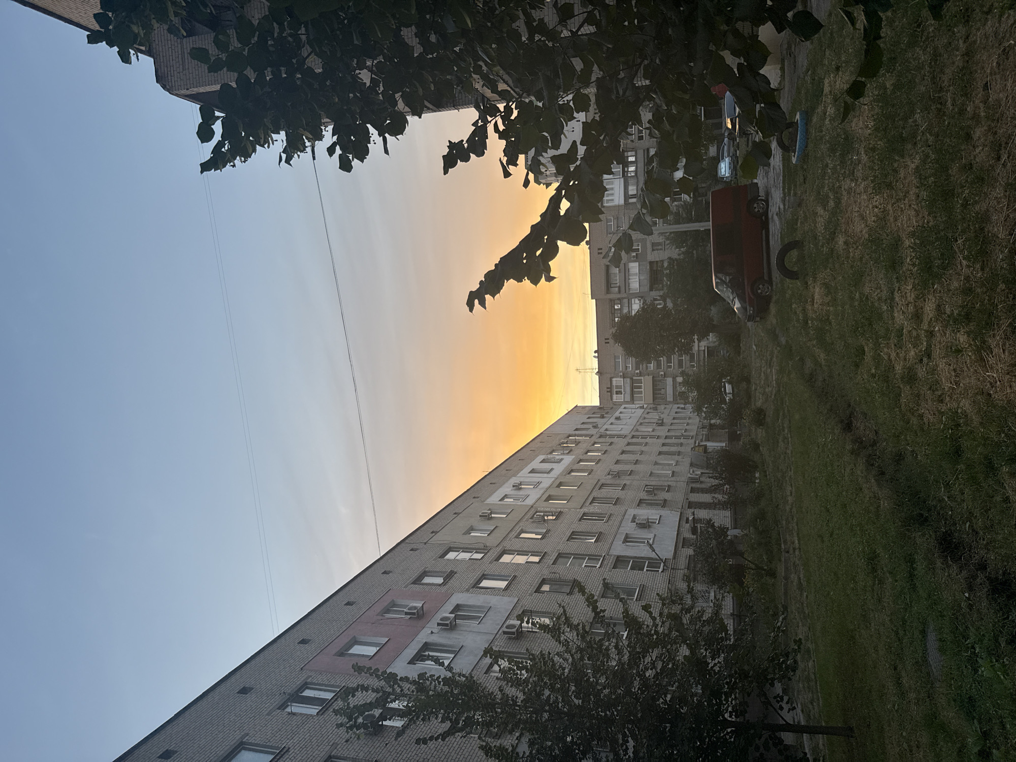 The image depicts a residential courtyard in Zaporizhzhia, Ukraine, during twilight. The scene is dominated by a long, multi-story brick apartment building on the left, with several windows and air conditioning units visible. A tree with dark green leaves stands in the foreground on the left, partially obscuring the building. The sky displays a gradient of colors, transitioning from a clear light blue at the top to a bright orange and yellow near the horizon, suggesting sunset or sunrise. In the middle ground, another apartment building is visible, with a red van parked in the courtyard. There is a small patch of dry grass in the courtyard, with some scattered trees and bushes. The overall impression is of a common urban residential area at dusk.