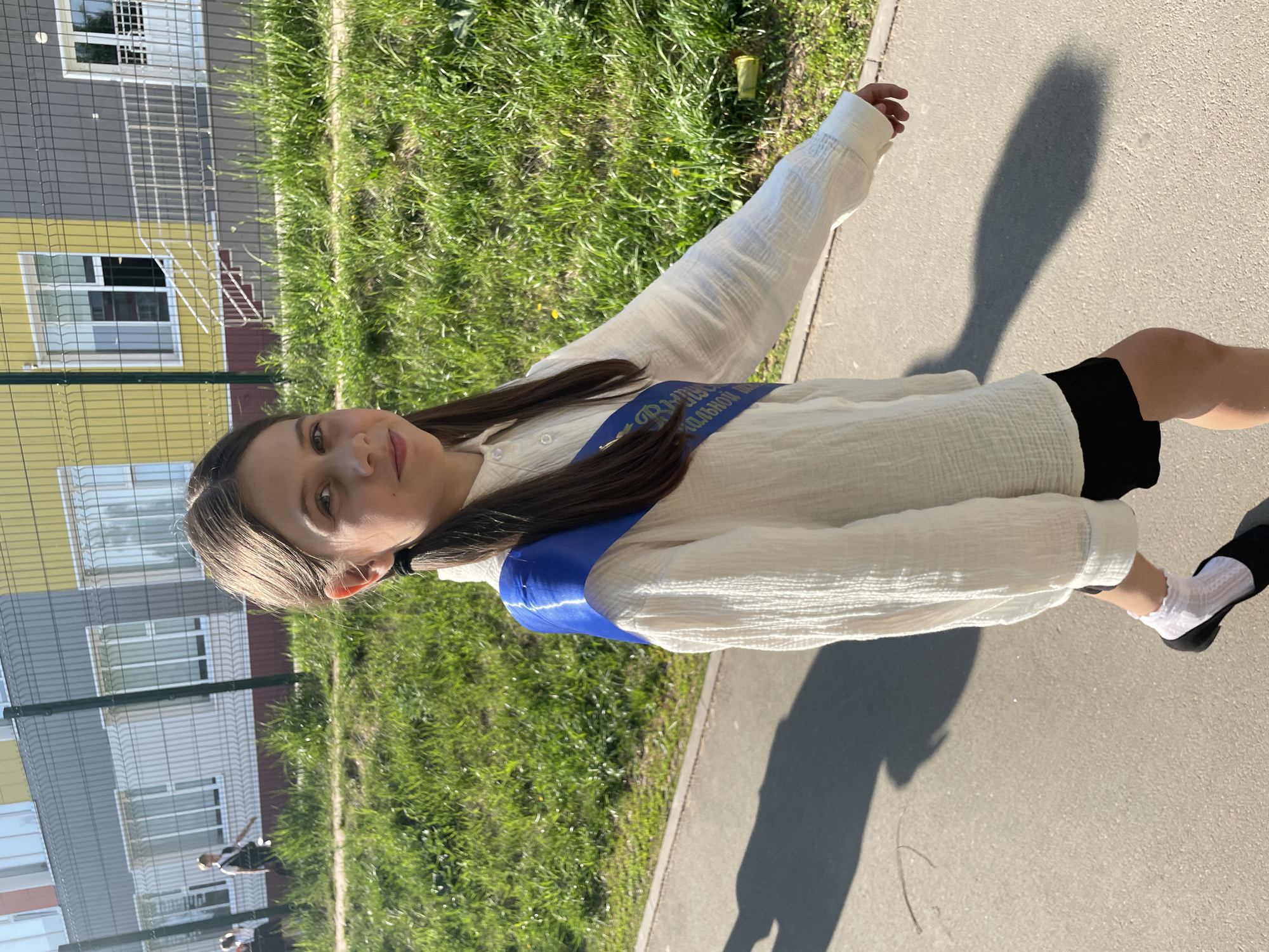 A girl wearing a white shirt and a blue sash stands outside. She also has on black shorts and black shoes with white ruffled socks. The background includes a fence, grass, and a building. There are other people in the distance. The location is Izhevsk, Russia.
