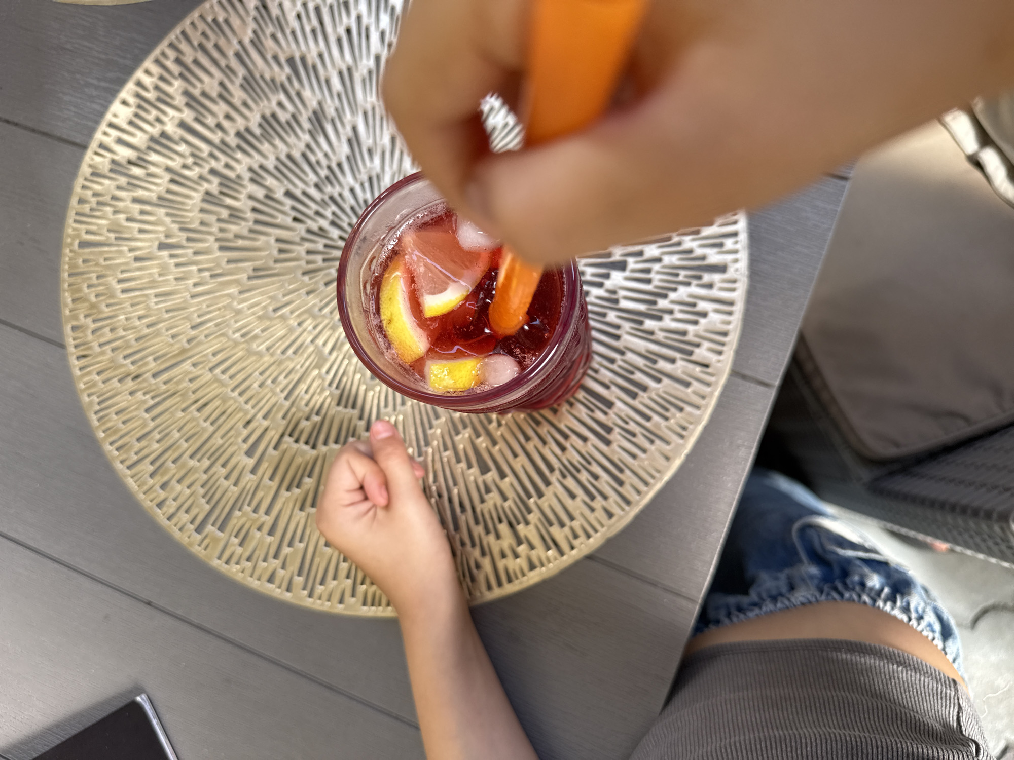 An overhead, close-up shot shows a child's left hand resting on a woven placemat next to a glass of red liquid with ice, lemon slices, and possibly berries. The child's right hand is visible in the foreground, holding an orange utensil and stirring the drink. The scene appears to be outdoors on a patio table. The table surface is gray, and a portion of the child's torso, wearing a grey and blue striped shirt and denim shorts, is visible at the bottom left. The background is blurred.