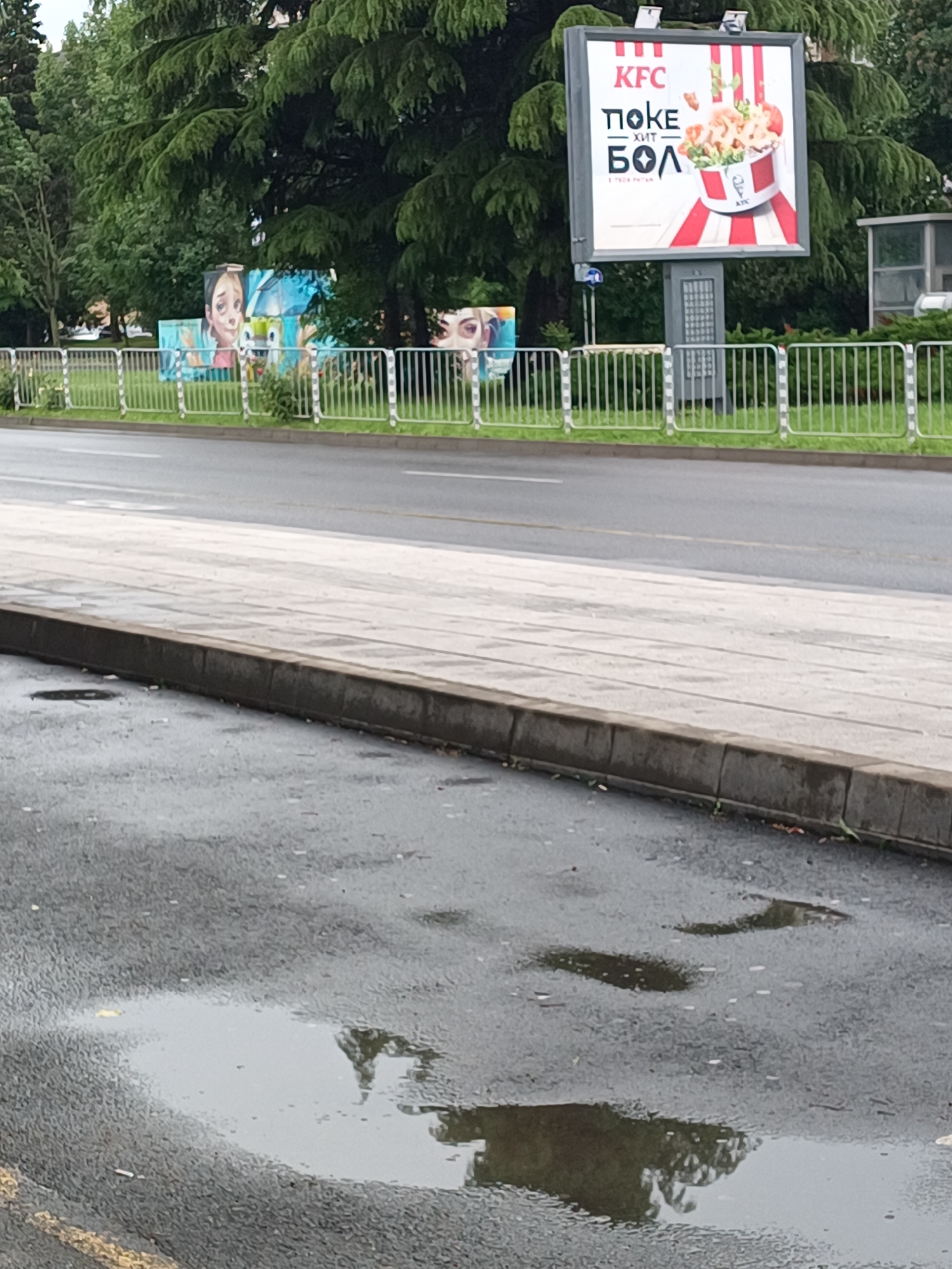 The image captures an outdoor street scene in Burgas, Bulgaria, seemingly after a rain shower, as evidenced by the wet asphalt road and several puddles in the foreground. A concrete curb separates the road from a paved sidewalk, beyond which a white metal fence borders a strip of green grass.
In the mid-ground, dense green trees form a lush backdrop. Two prominent visual elements stand out against the greenery. On the right, a large digital billboard advertises KFC, displaying an image of a bucket filled with food. The text on the billboard is in Cyrillic, reading "KFC" and "ПОКЕ ХИТ БОЛ," which translates to "POKE HIT BOWL." To the left, behind the fence and partly obscured by trees, vibrant street art or murals are visible. One section clearly depicts a stylized face with large, expressive eyes, and another partial face can be seen further to the right.
The time of day appears to be daytime, likely overcast, given the diffused lighting and lack of strong shadows. The puddles on the wet road reflect the surrounding trees, adding a subtle visual dynamic to the quiet urban setting. No people are present, and no specific activity is taking place, presenting a still snapshot of the street.