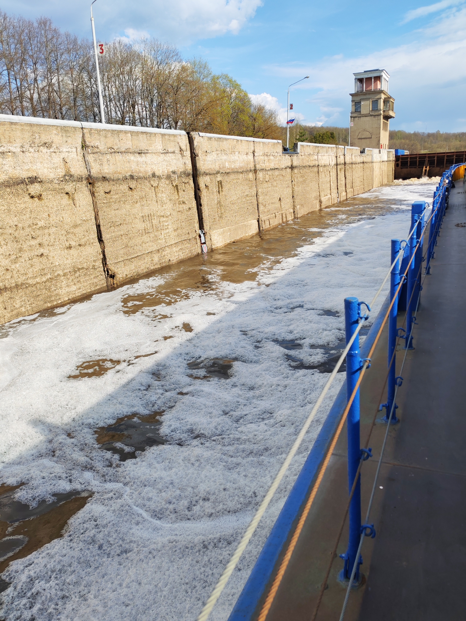 The image shows a lock channel filled with frothy water, likely a result of turbulence from water being released or pumped through. The channel is defined by high, concrete walls that show signs of age and weathering. On the right side of the frame is a blue railing, indicating a pedestrian or viewing area. In the background, a control tower is visible, with a distinctive boxy architectural style. The Russian flag flies in the background. Bare trees and some vegetation are visible beyond the tower. Based on the tower style, flag, and structures, the location is plausibly in Yakhroma, Russia, near a canal or river lock.