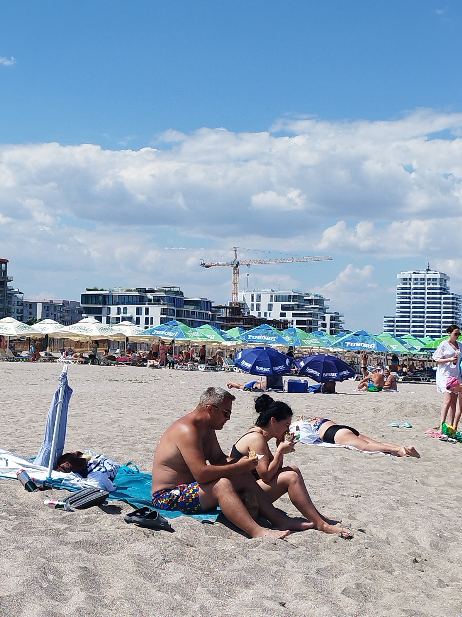 This image depicts a crowded beach scene in Constanța, Romania, on a sunny day with scattered clouds. In the foreground, a man and a woman are seated on the sand. The man, wearing sunglasses and colorful swim trunks, is looking to his right. The woman, with dark hair pulled up, is wearing a bikini and eating. To their left, a dog is resting on a blue towel, with a beach umbrella partially obscuring it. Several pairs of flip-flops and discarded cans are scattered on the sand.
In the background, a row of beach umbrellas, some displaying the "Tuborg" logo, provides shade for beachgoers lounging on chairs. Further back, modern buildings and construction cranes indicate the urban development of the area. A tall white building with many windows is visible on the right. Several individuals are walking and relaxing on the beach. The overall scene suggests a typical summer day at a popular coastal resort.