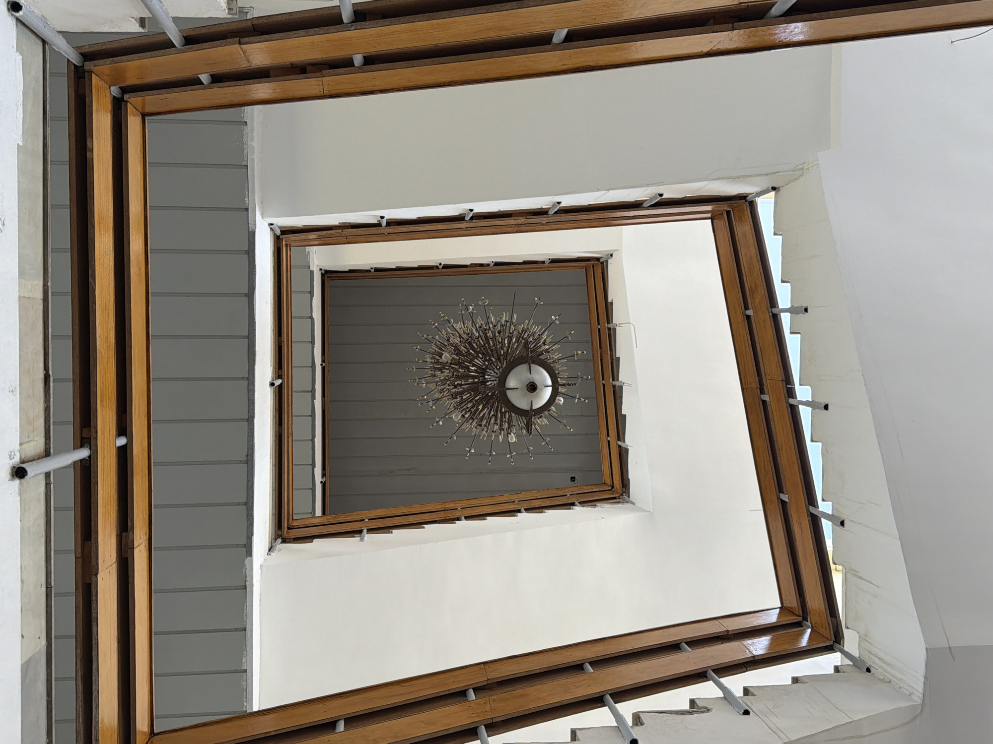 The image, captured from a low angle looking upwards within a building, shows a square skylight or ceiling opening framed by dark wood. Below this opening, a metallic chandelier with a sunburst design and visible bulbs hangs centrally. The structure appears to be a stairwell, with white walls and stairs visible to the sides, bordered by wooden railings and metal supports. The ceiling above the wooden frame is textured and grey. The location, Changi, Uzbekistan, is mentioned.