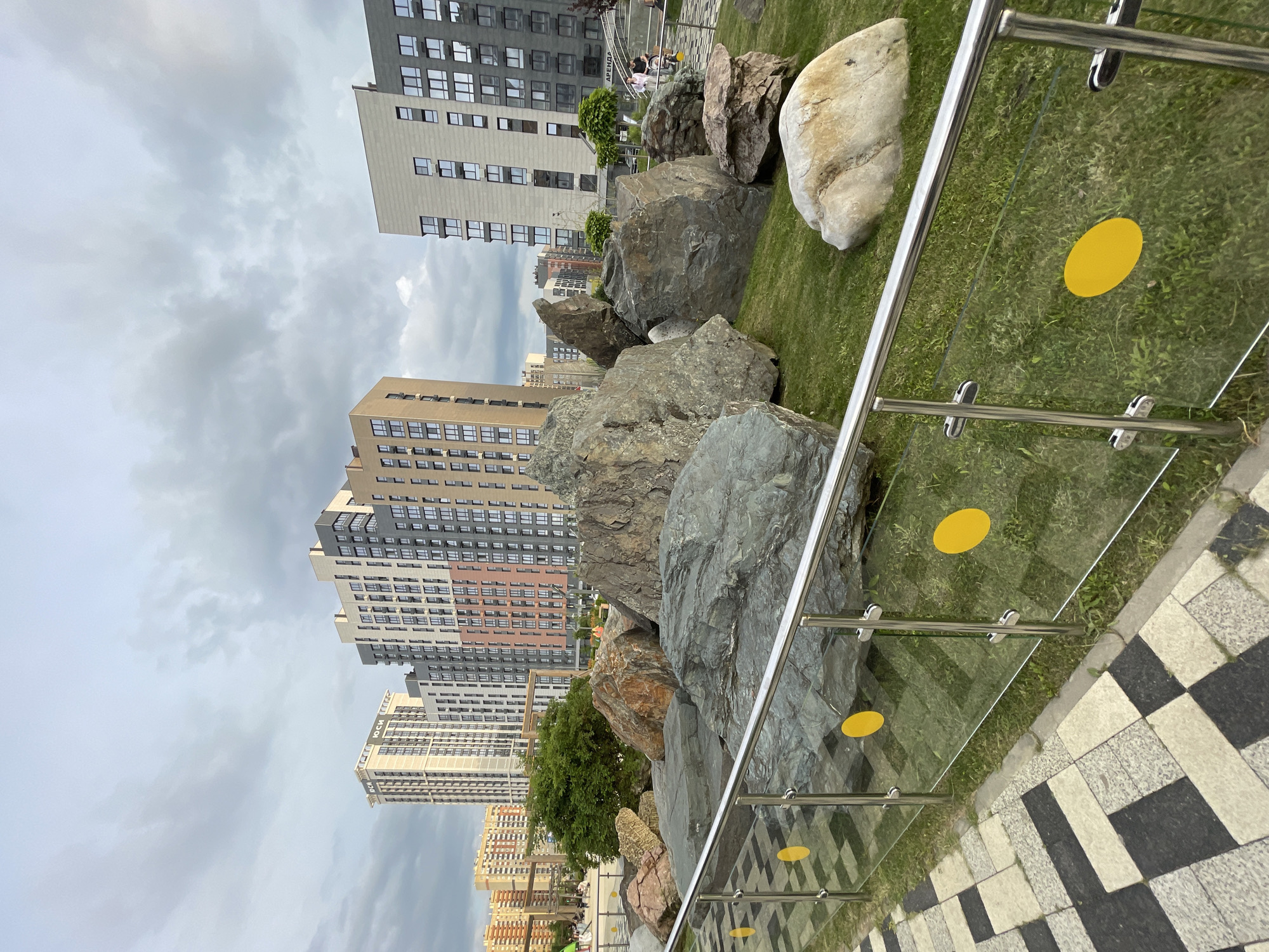 This media file depicts an outdoor scene with a focus on a landscaped area featuring large rocks and a modern railing system. In the background, several tall, multi-story apartment buildings are visible, suggesting an urban or suburban setting. The buildings have a contemporary architectural style with numerous windows.
The foreground is dominated by a collection of substantial rocks of varying sizes and colors, scattered across a green, grassy area. A polished metal railing with glass panels runs along the edge of the landscaped area. Yellow circular decals are present on some of the glass panels. The ground beside the landscaped area is paved with a checkered pattern of light and dark gray tiles.
While the image is largely focused on the landscape and architecture, there are glimpses of activity in the background. A few people can be seen walking in the distance, near the buildings and the landscaped rocks, indicating a public space. One of the crops shows a sign with Cyrillic text, likely related to a business or area name, reading "АРЕНДА" which translates to "RENT".
The sky is overcast with gray clouds, suggesting a cloudy or possibly impending rainy day. The lighting is diffused, indicating it's likely daytime, but without direct sunlight. The overall mood is calm and perhaps a bit somber due to the weather. The location is identified as Stavropol, Russia, which helps contextualize the architectural style and the presence of Cyrillic text.
