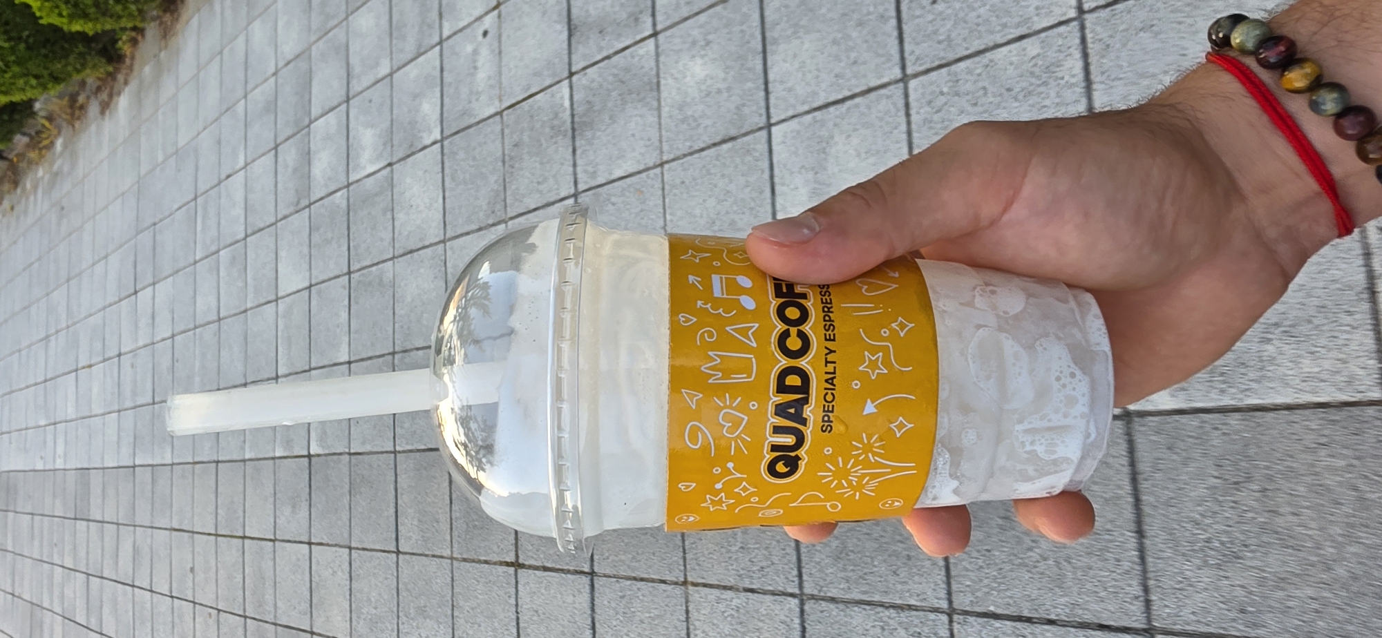 An overhead shot shows a person's hand holding a large, clear plastic cup filled with a frothy, white beverage, likely a coffee drink. The cup is adorned with a bright yellow paper sleeve featuring playful graphics and the text "QUAD COF" followed by "SPECIALTY ESPRESSO". A clear plastic lid covers the cup, and a white plastic straw extends from it. The cup shows condensation on the outside, indicating it's a cold drink. The background is a paved walkway composed of square, grey tiles. To the right, a blurred green bush can be seen. The image was captured in Cheongju-si, South Korea.