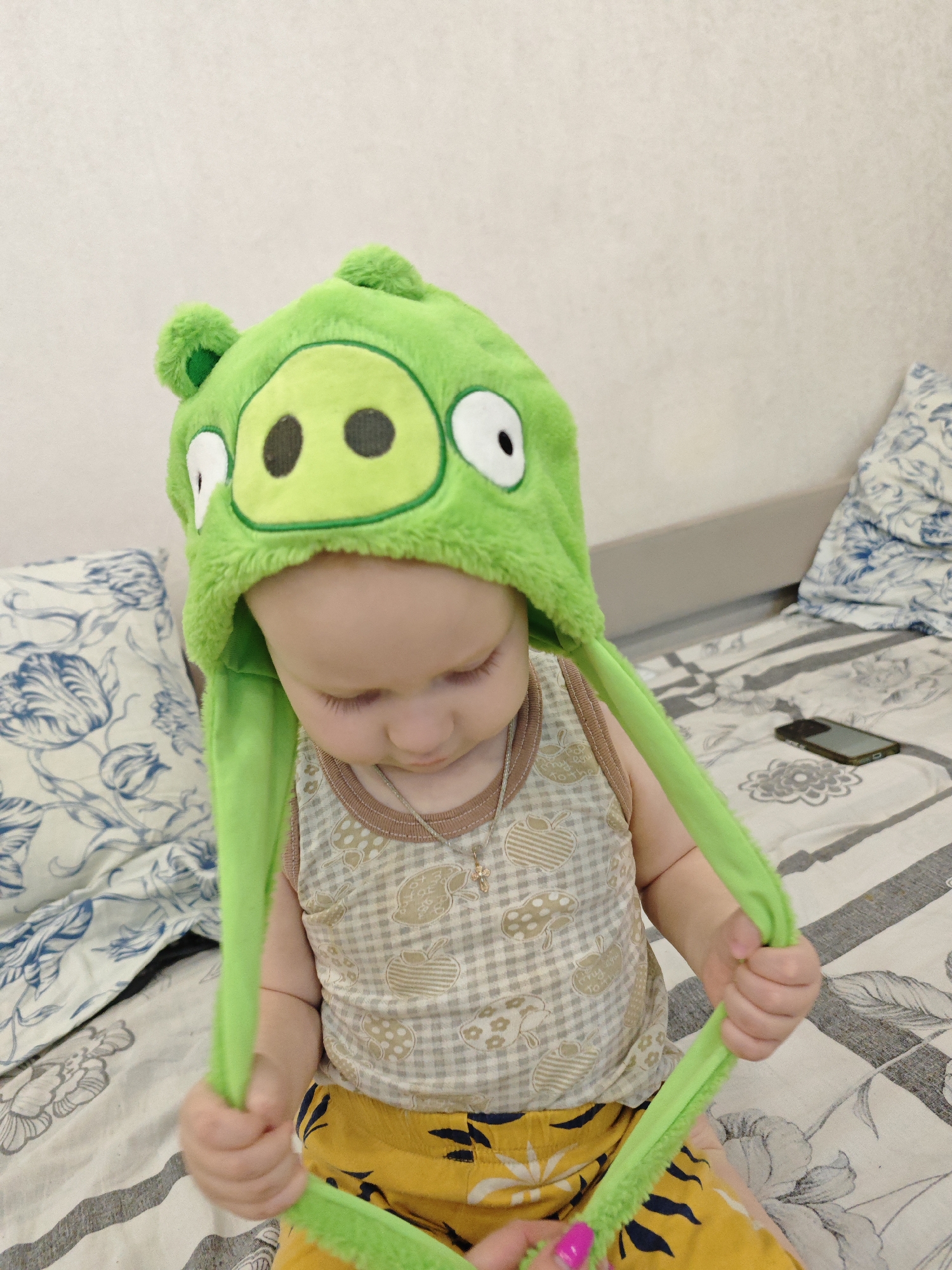 A toddler is sitting on a bed. The toddler is wearing a green pig hat, a beige patterned tank top, and yellow shorts with a leaf pattern. The toddler is holding the straps of the hat in both hands. A smartphone is on the bed. The bedding has a floral pattern. The location is Markovskii, Russia.